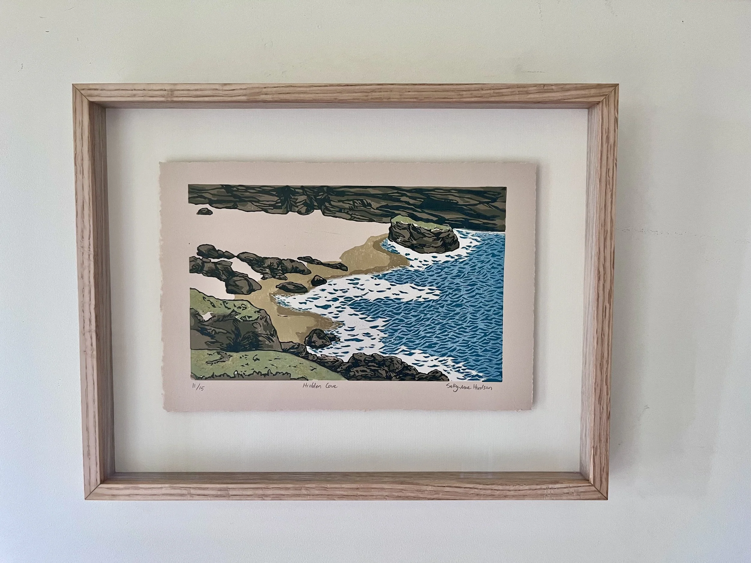A float framed artwork titled 'Hidden Cove' by Sally Mae Hudson, numbered 11/15, depicting a coastal scene with rocks, a sandy beach, and ocean waves.