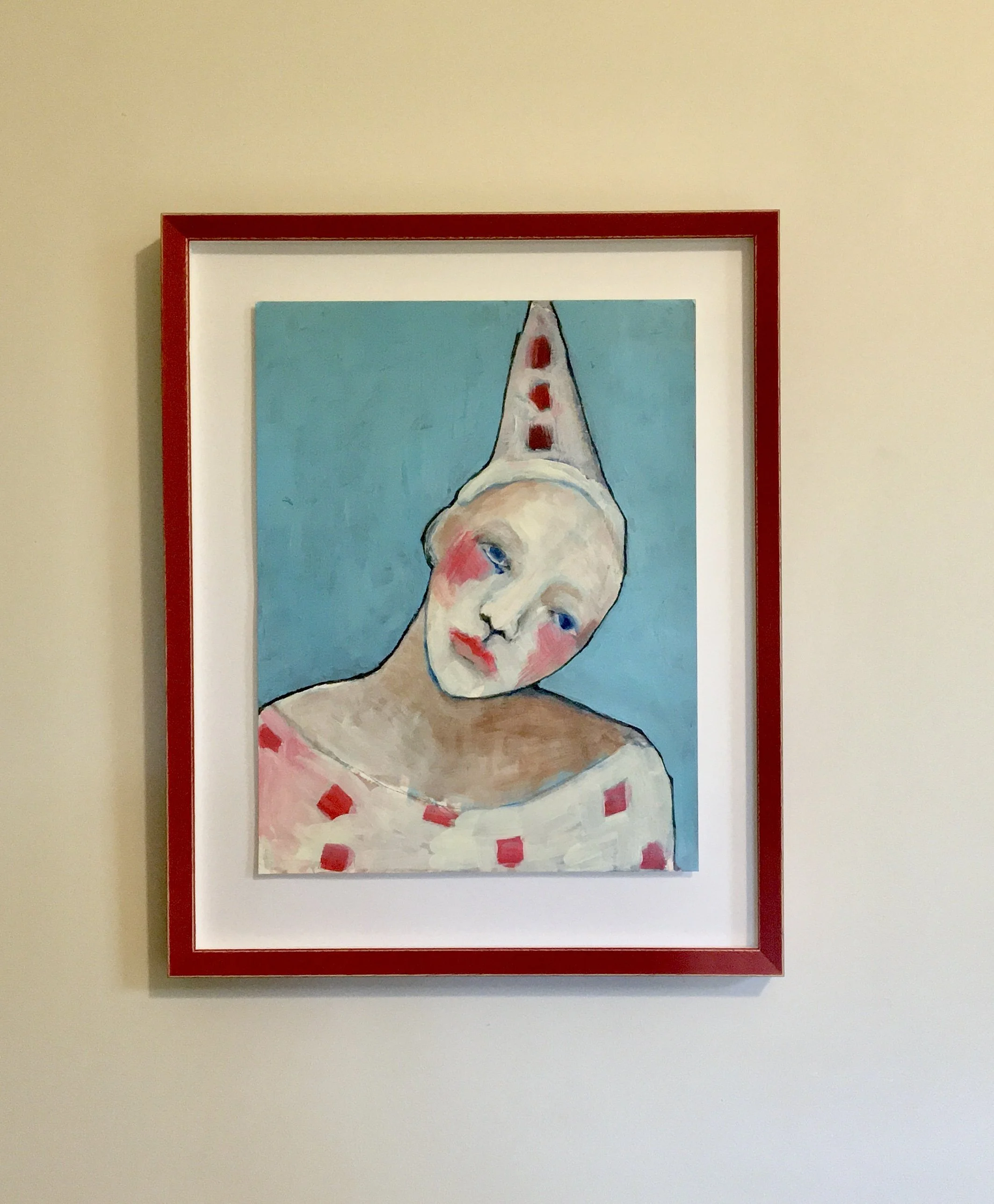A framed painting of a clown in a hat with a clinched left eye, wearing a white dress with red squares, against a blue background, mounted on a beige wall.