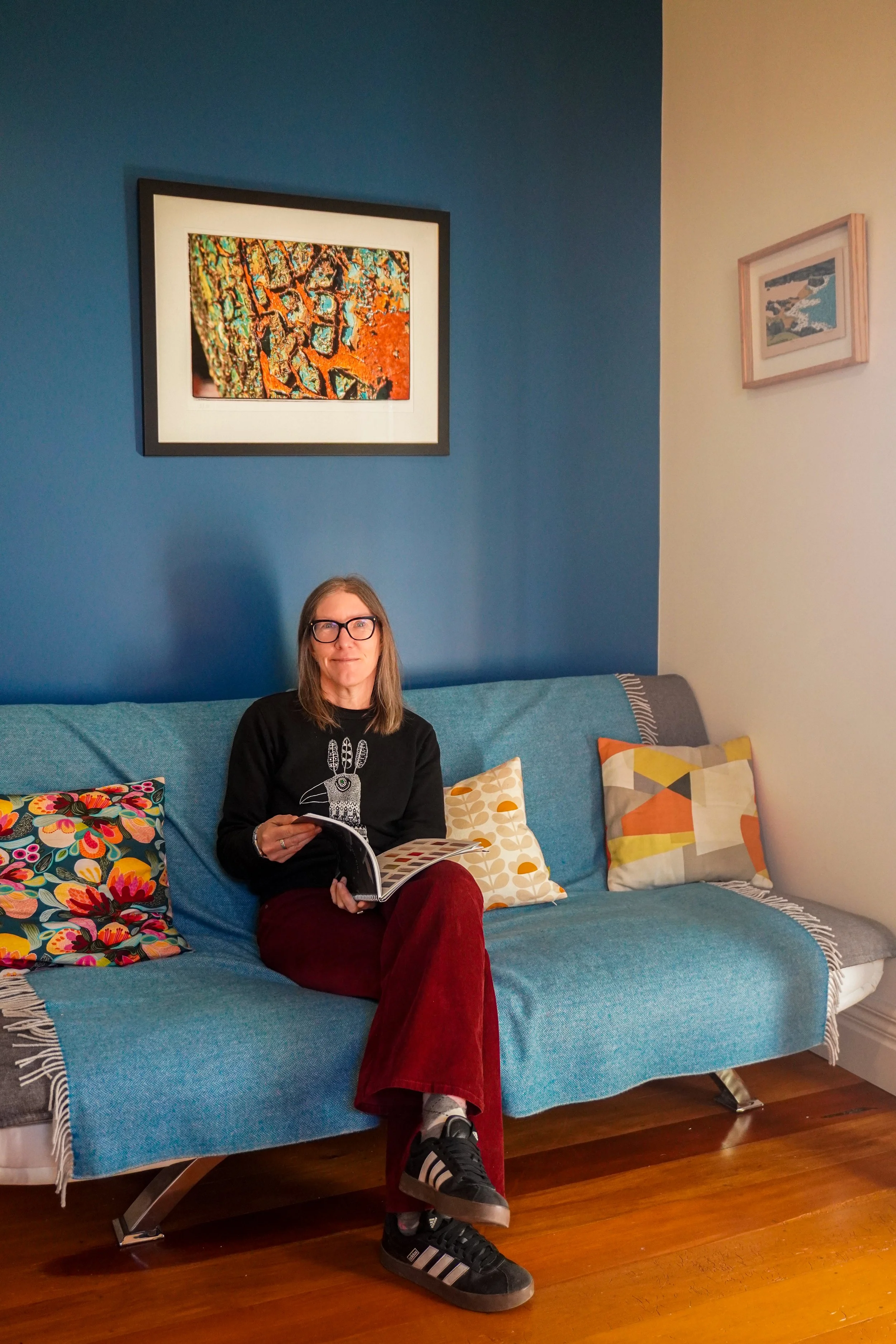 A woman with glasses sitting on a blue sofa with colorful patterned pillows, holding a magazine, in a room with blue and cream walls decorated with framed artwork.