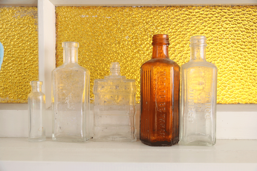 Five antique glass bottles of various sizes and shapes, with some having embossed text, displayed on a shelf in front of a textured yellow background.