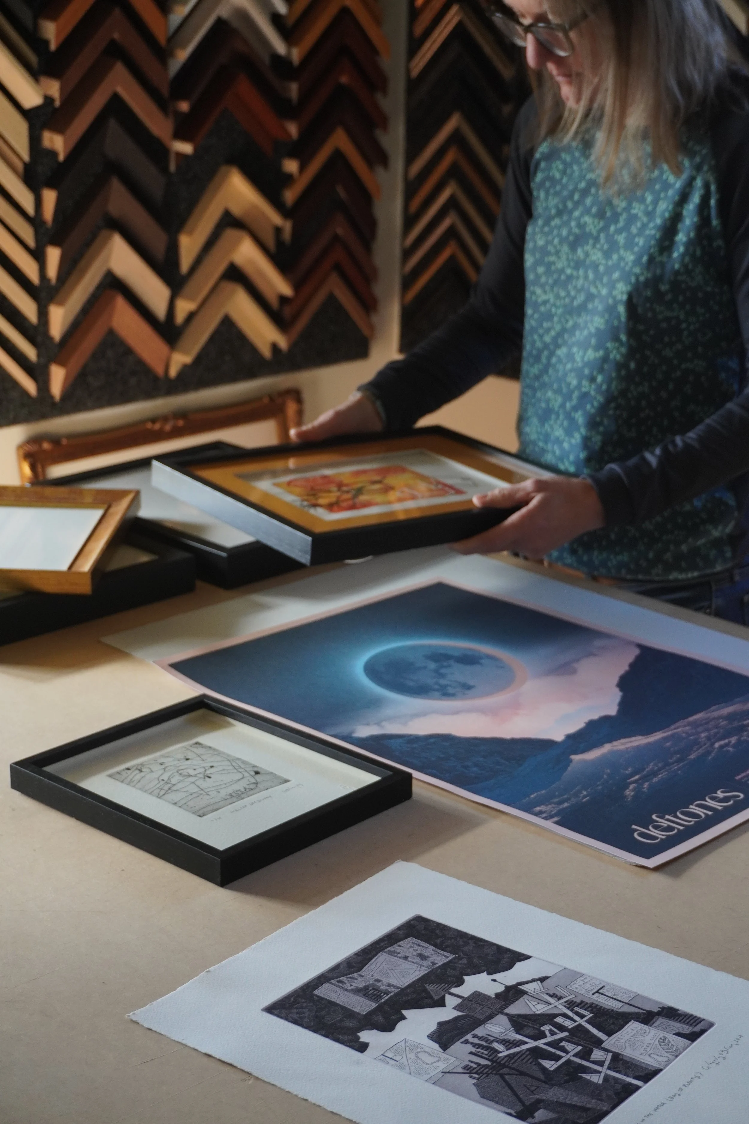 A woman with long hair and glasses holds a framed artwork in an art gallery or store. Several framed artworks and prints are laid out on a table in front of her, with a wall of framed picture corners in the background.