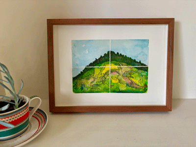Framed watercolor painting of a green hill under a blue sky, divided into four sections, leaning against a white wall next to a ceramic plant pot with a plant.