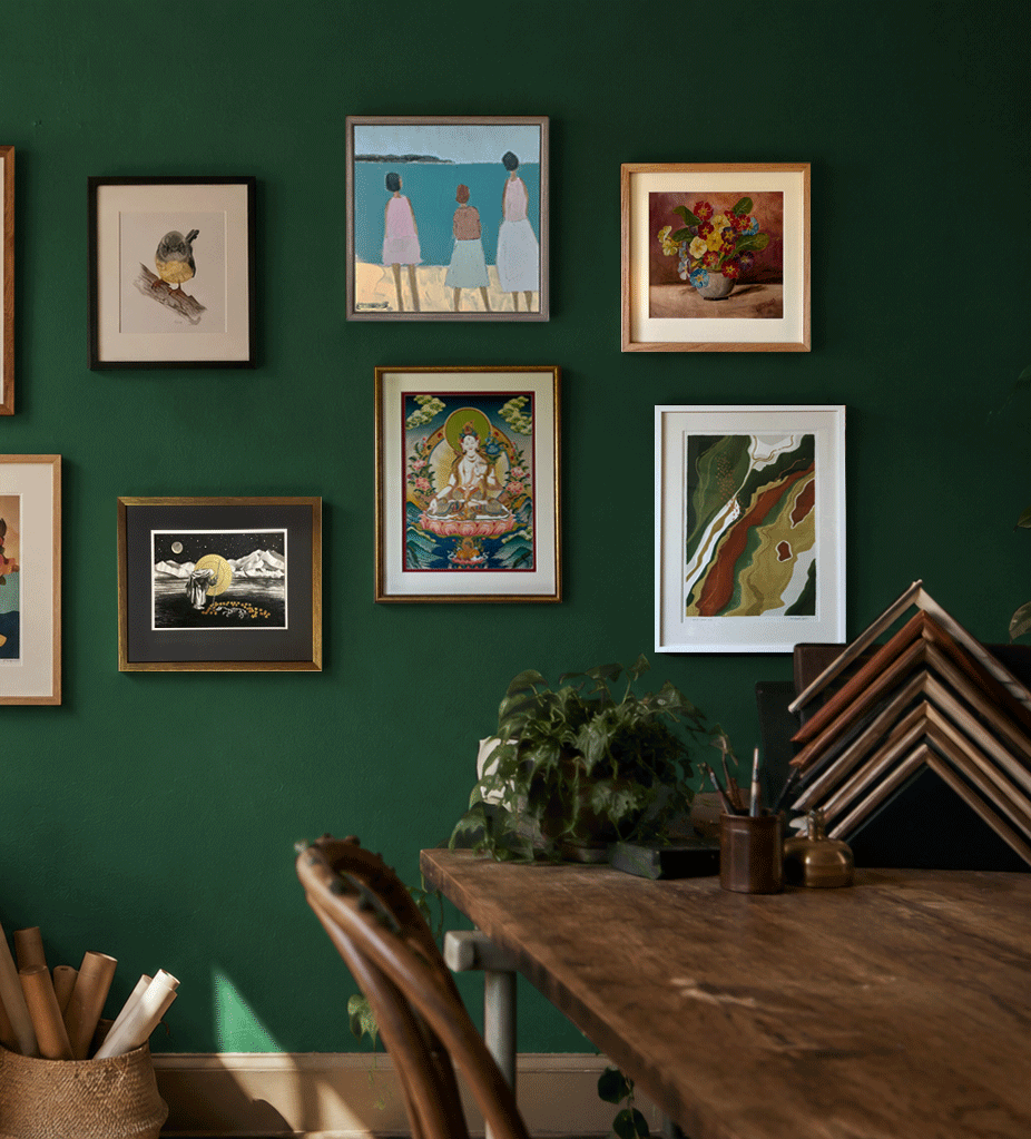 Indoor space with green wall decorated with various framed artworks, including a bird, people at the beach, a floral still life, a celestial scene, a deity, and abstract art. Part of a wooden table with green plants, picture frames, and art supplies is visible in the foreground.