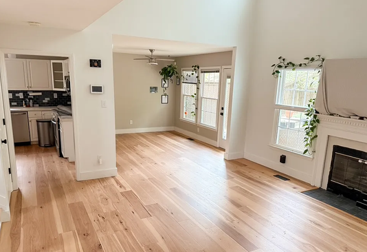 Empty living room with hardwood floors, white walls, large windows, and a fireplace with decorative greenery above it.