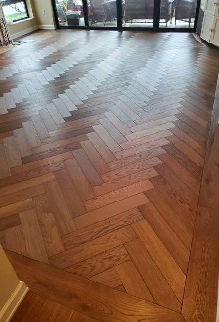 Close-up view of a wooden floor with a herringbone pattern in a room with large windows, natural light, and sliding glass doors.