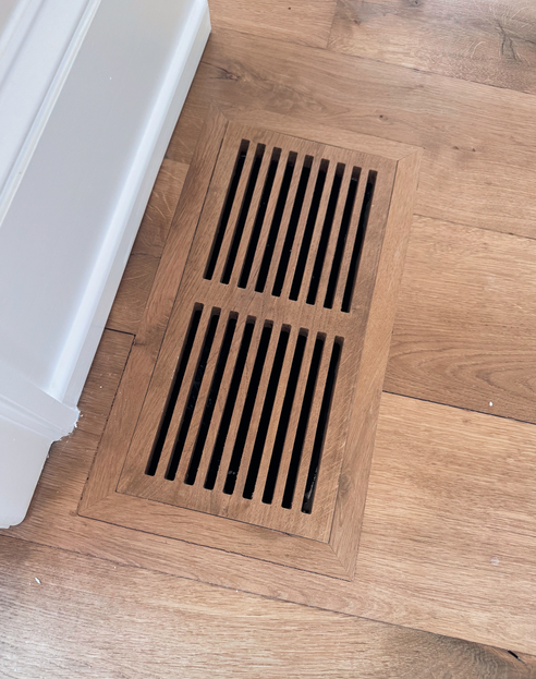 Wooden floor vent cover with black grille slats in a house interior.