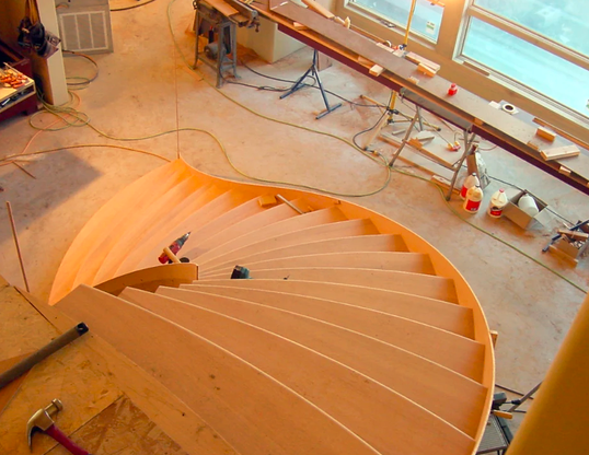 View of a wooden spiral staircase under construction in a room with large windows, construction tools and supplies nearby.