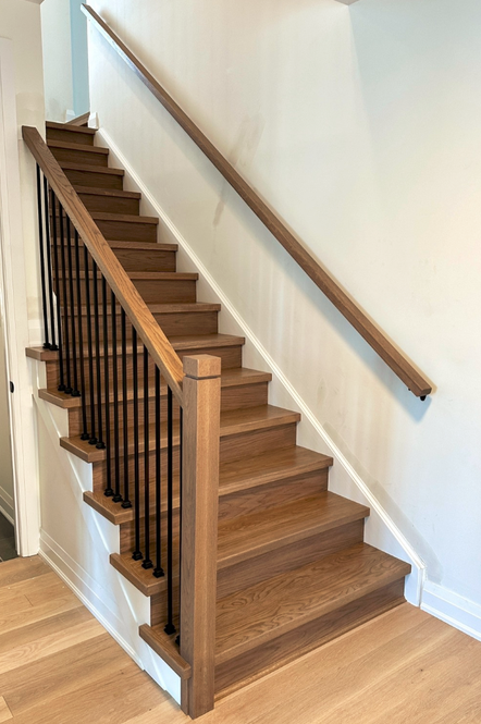 Wood staircase with black metal balusters and handrail, white wall, and wooden flooring.