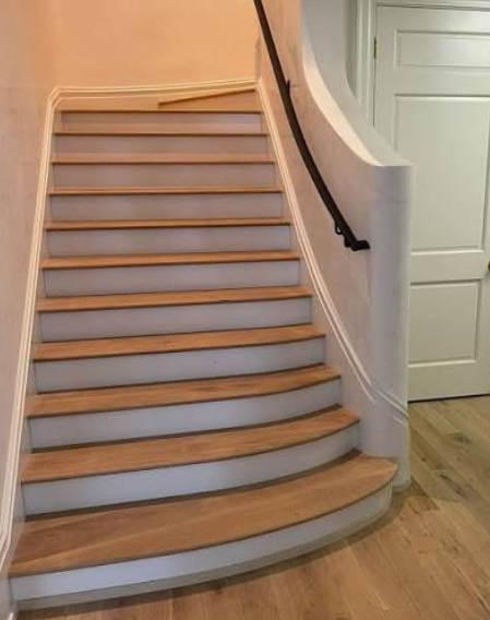 Curved staircase with wooden steps, white risers, black handrail, and white walls. A door is visible at the top of the stairs.