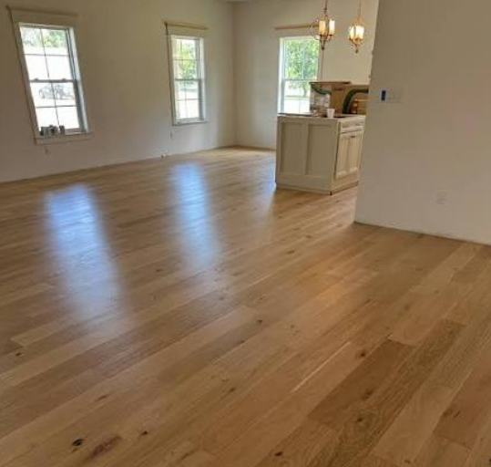 Empty room with hardwood floors, three windows, and a kitchen counter with hanging lights.
