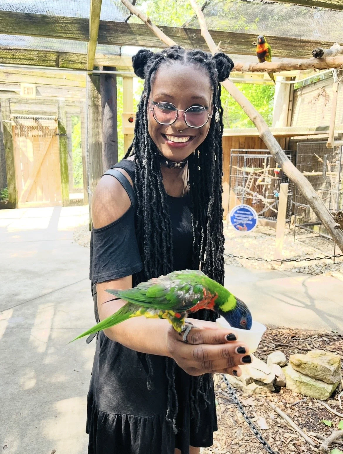 A woman with glasses and dreadlocks smiling while holding a colorful bird in her hand at a bird sanctuary.