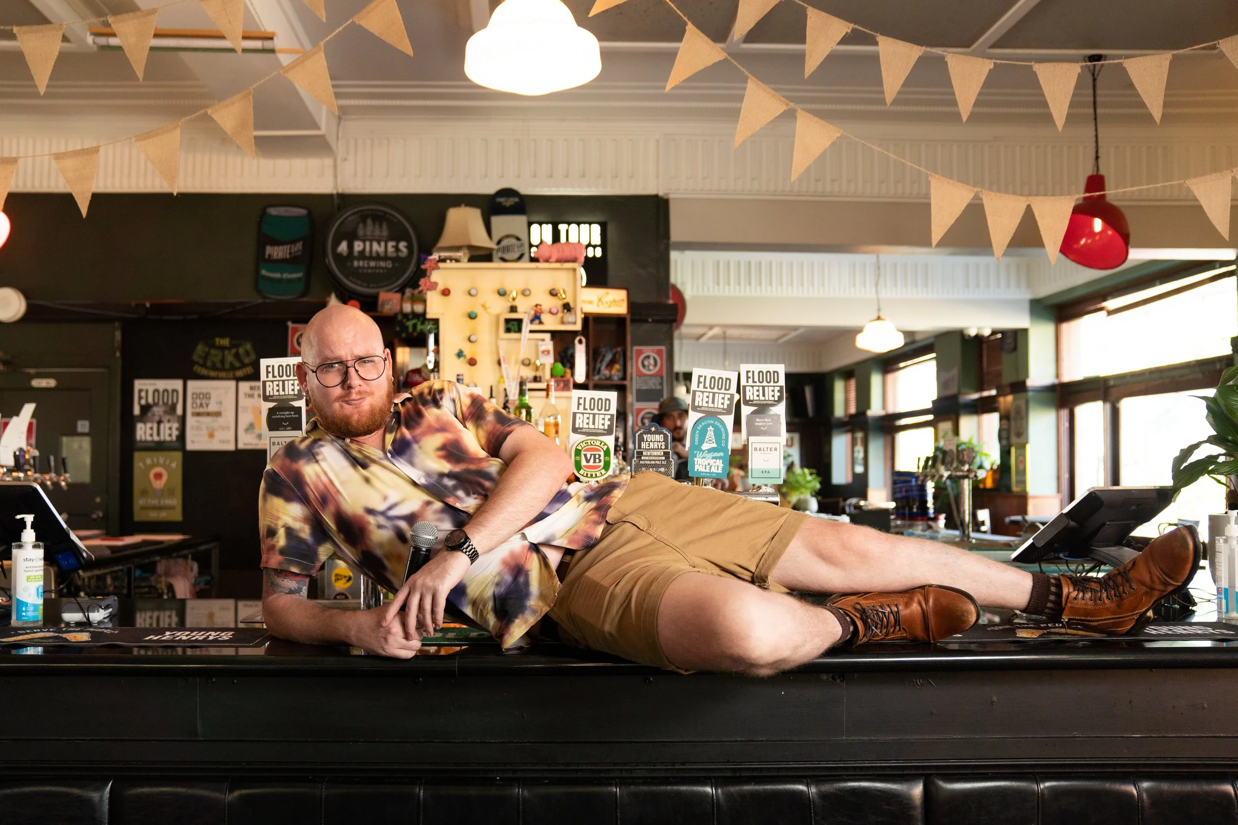 A person with glasses and tattoos holding a microphone, sitting at a wooden table with a black bottle labeled 'RESERVE' and surrounded by many Polaroid photos of dogs on the wall behind him.