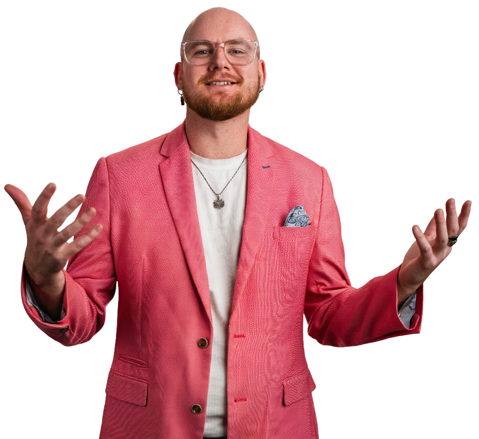A man with glasses, a beard, and a bald head, wearing a pink blazer over a white shirt, with a necklace and pocket square, smiling with one hand raised in a gesture.
