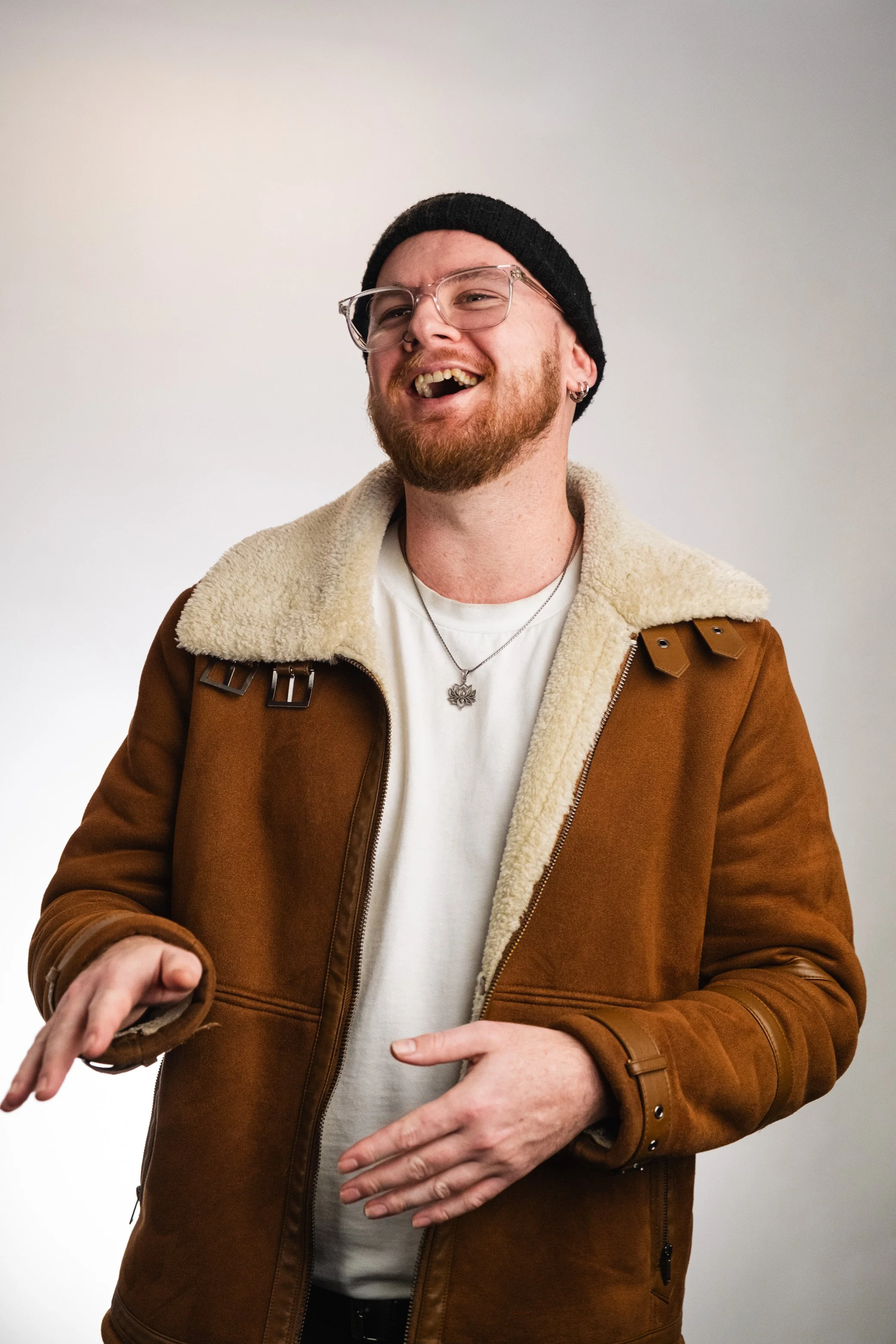 A man with a beard and glasses laughing, wearing a black beanie, a white shirt, and a brown jacket with a shearling collar, standing against a plain white wall.