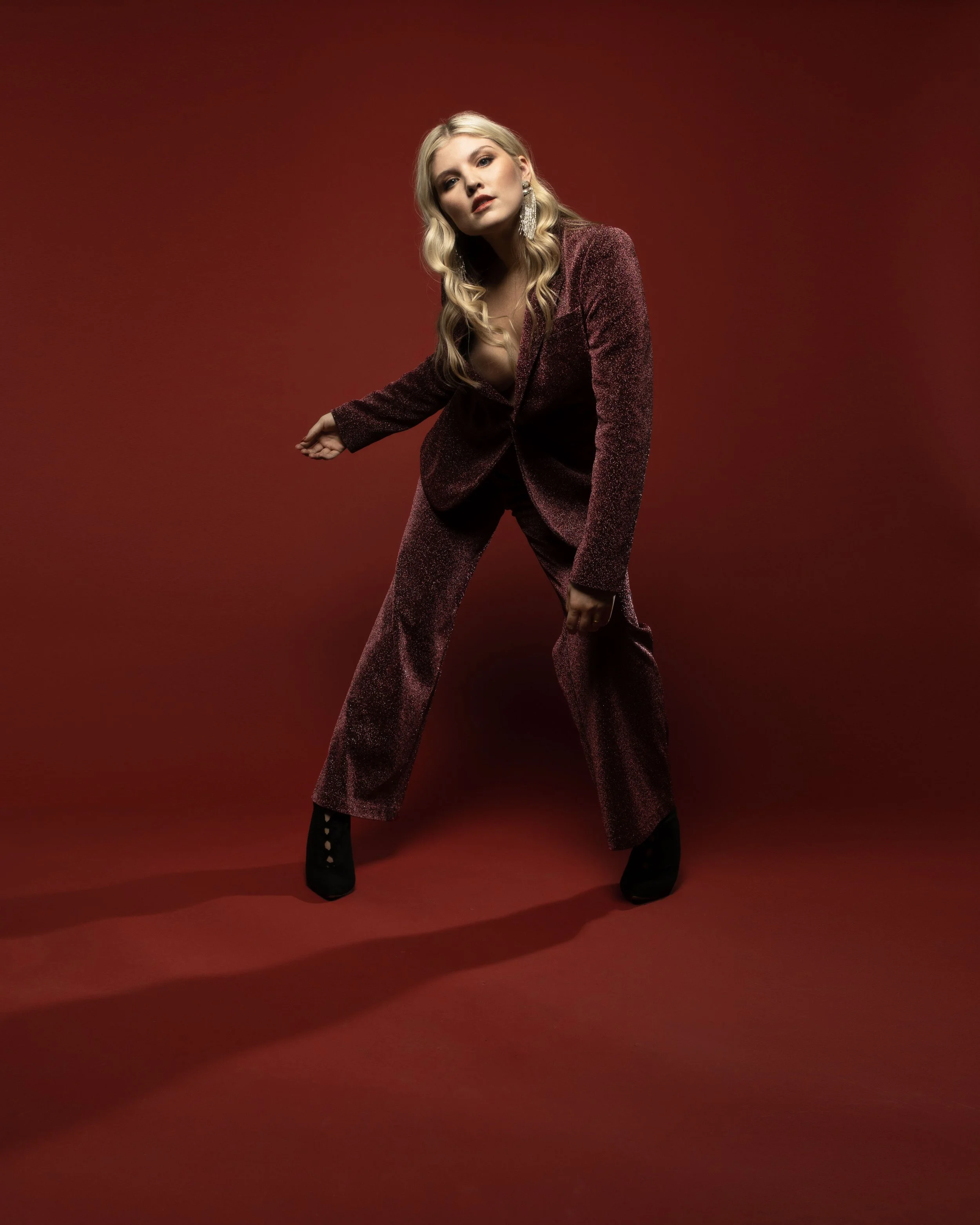 A woman with long blonde hair styled in loose waves, wearing a glittery burgundy blazer and matching wide-leg pants, poses against a dark red background. She has dramatic makeup, large dangling earrings, and black high-heeled boots, creating a glamor