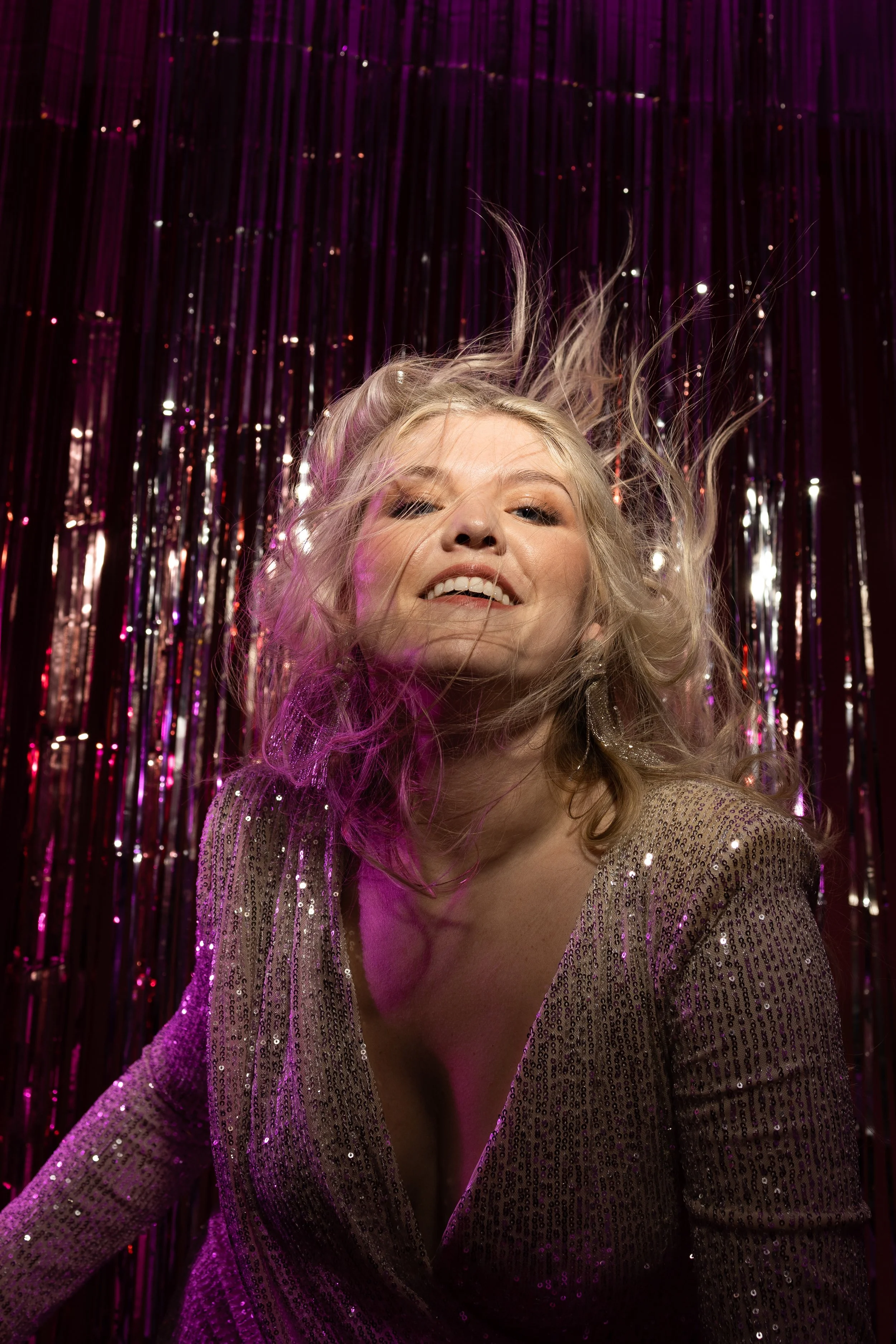 A woman with curly blonde hair smiling against a purple sequin backdrop, wearing a glittery, gold, low-cut dress.