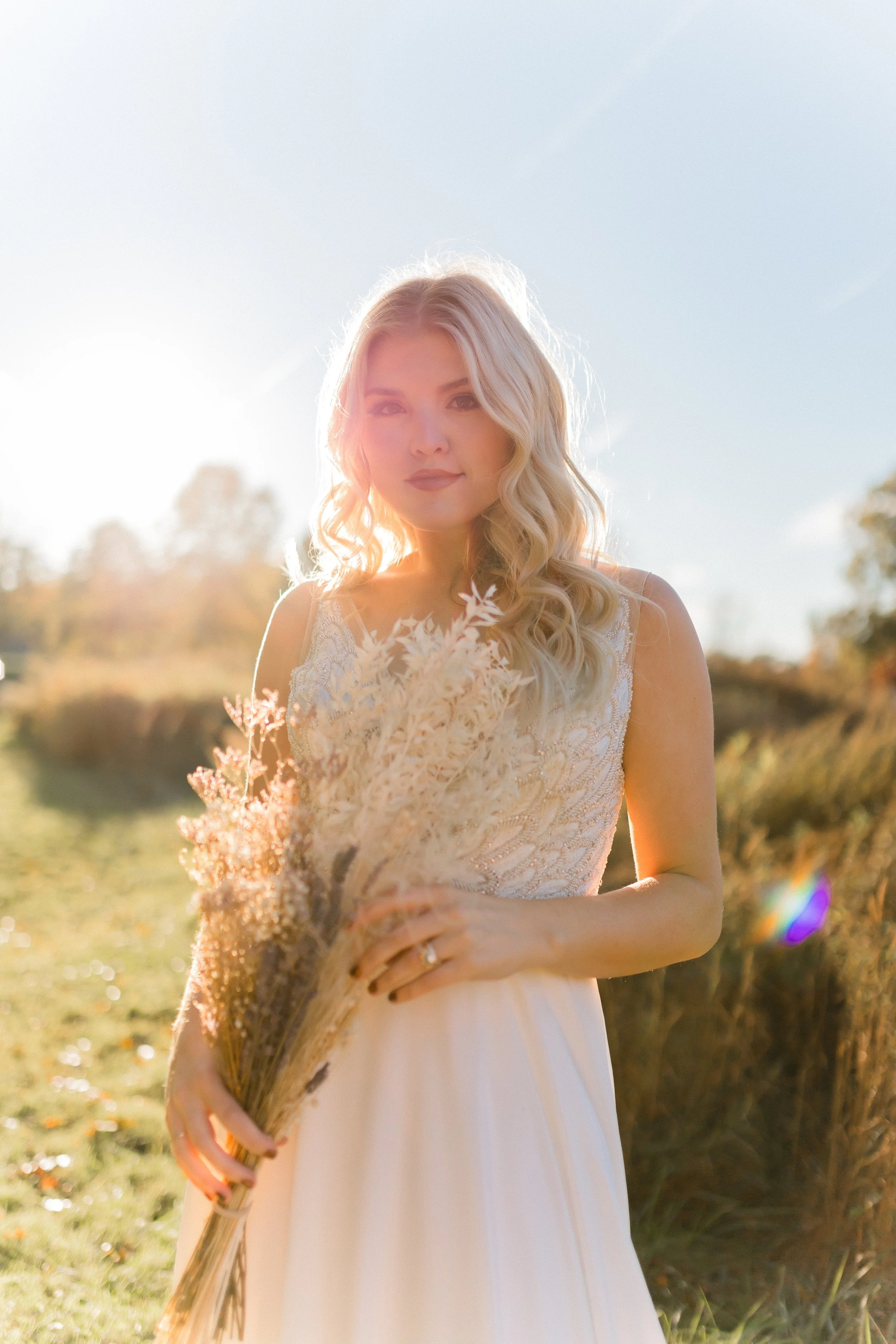 A woman with long blonde hair in a white lace dress holding a bouquet of dried flowers outdoors in sunlight.