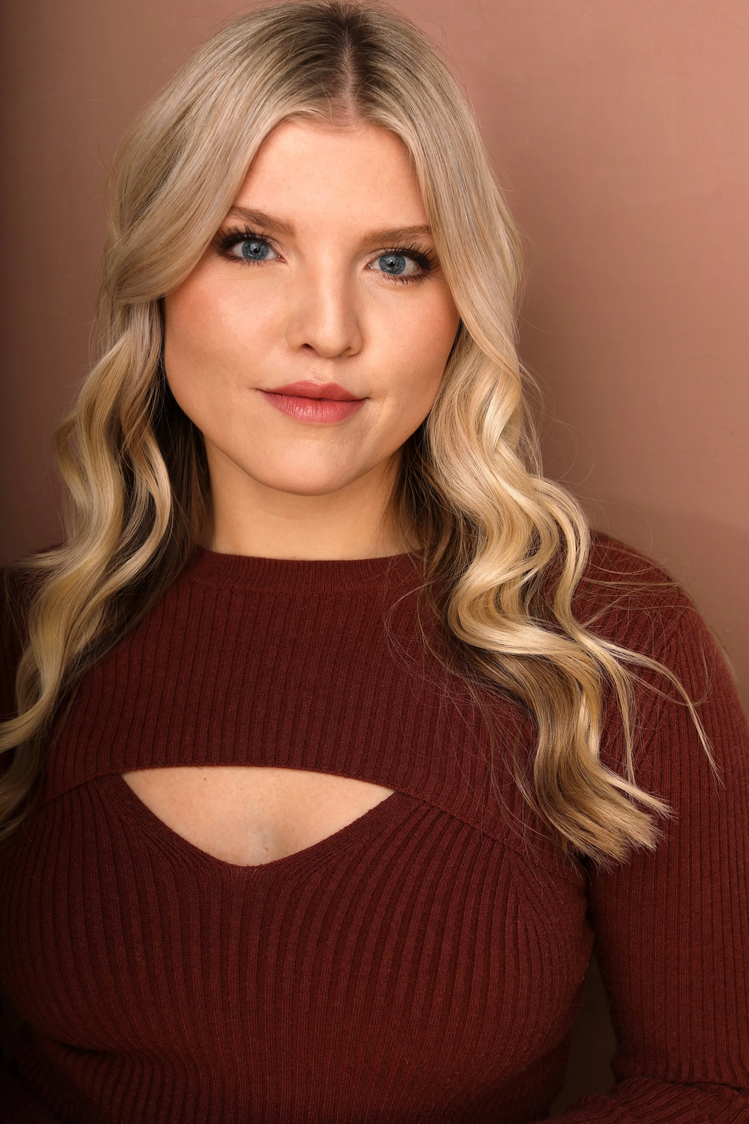 Portrait of a young woman with long blonde wavy hair, blue eyes, wearing a burgundy shirt with a cutout detail on the chest, standing against a plain background.