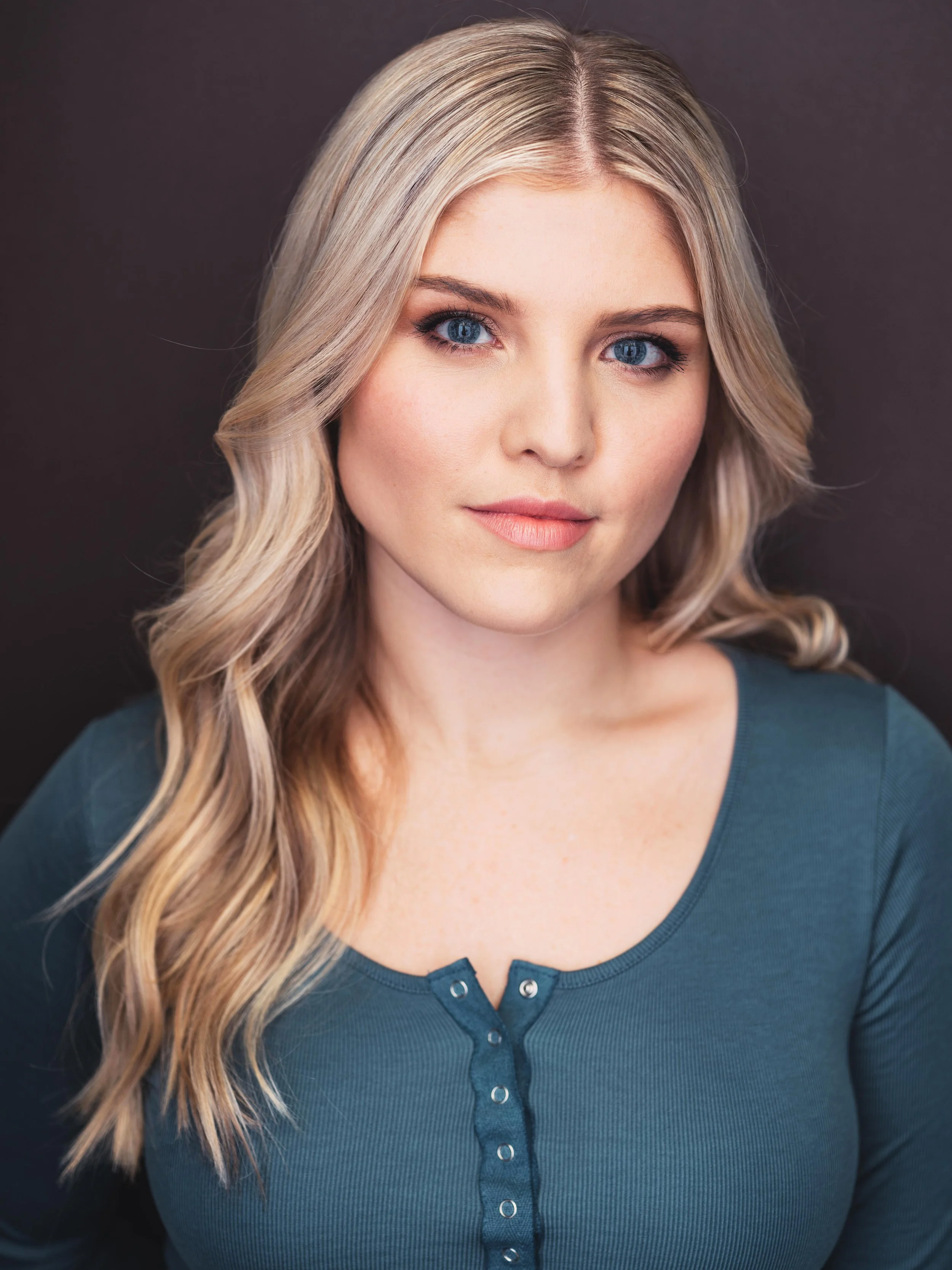 Close-up of a young woman with long blonde hair styled in loose waves, wearing a teal buttoned shirt, looking straight at the camera against a dark background.