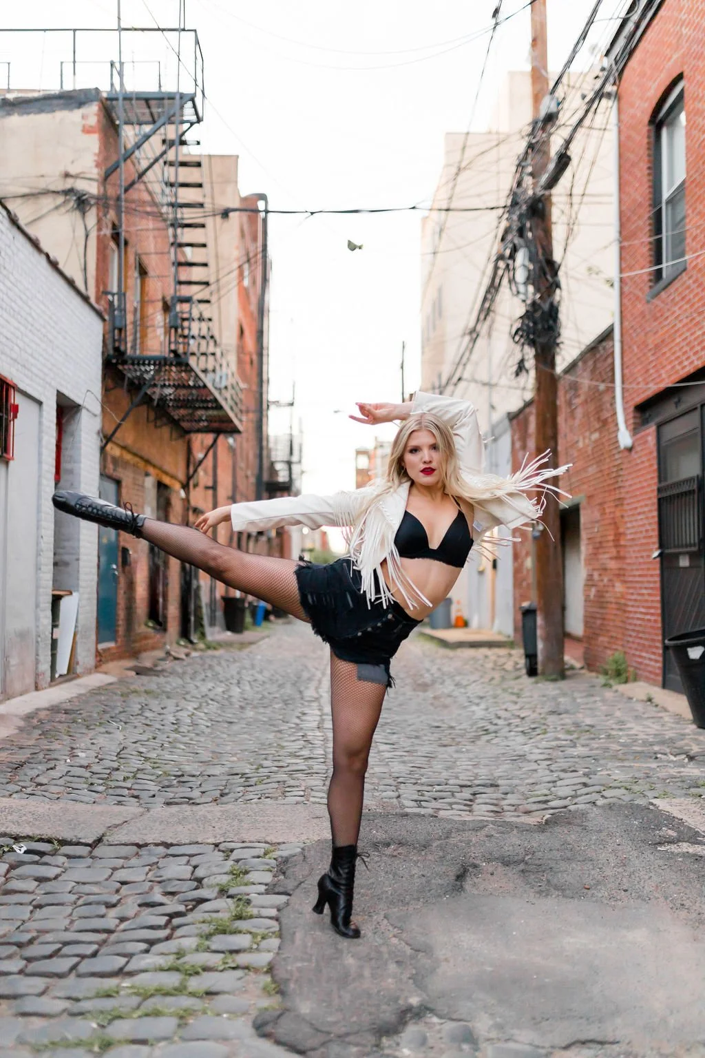 A woman with blonde hair in a dance pose in an alleyway, wearing a black bra, black shorts, fishnet stockings, high heel boots, and a white fringed jacket.