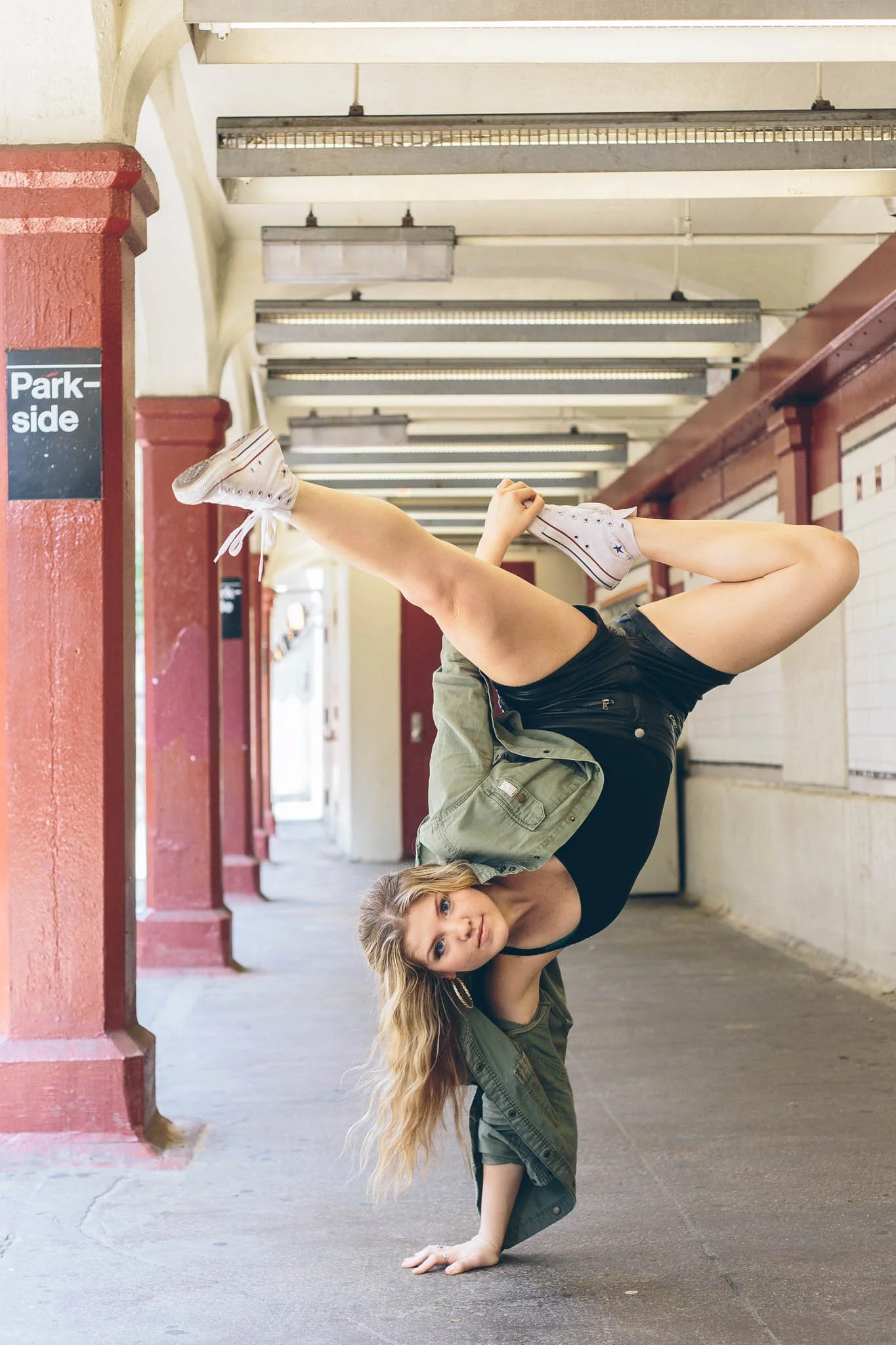 A woman in black top and green jacket performing a handstand with a young girl in a green jacket and white sneakers balancing on her shoulders in a covered outdoor corridor with red brick columns and fluorescent lighting.