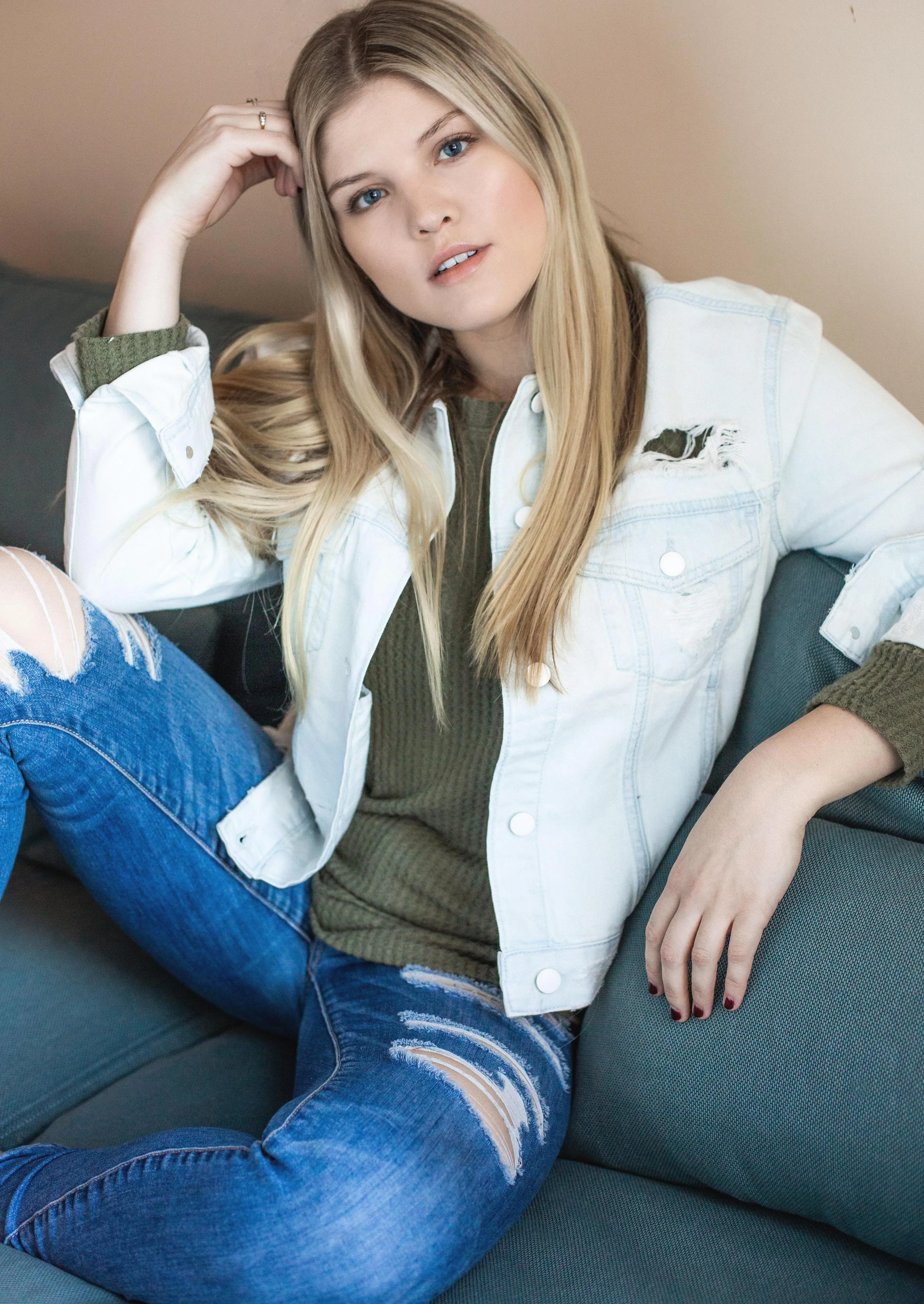 Young woman with long blonde hair, blue eyes, and fair skin, sitting on a gray couch, wearing a white denim jacket with a ripped shoulder, green top, and ripped blue jeans.