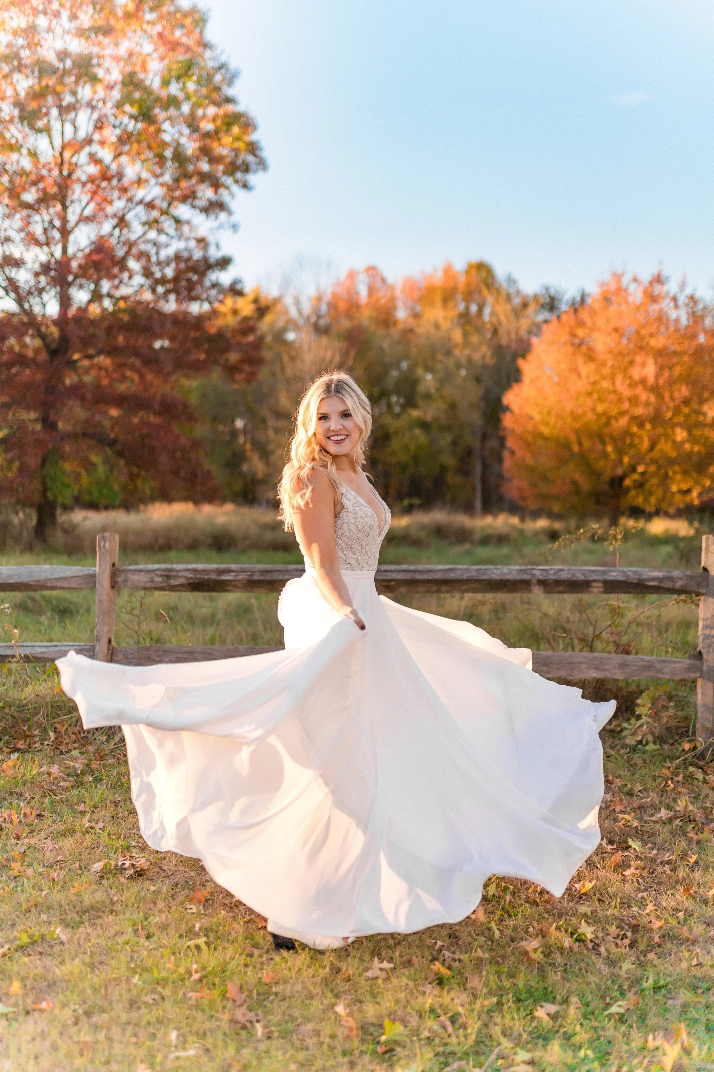 A woman in a white wedding dress standing outdoors in a park with autumn foliage, smiling and twirling her dress.