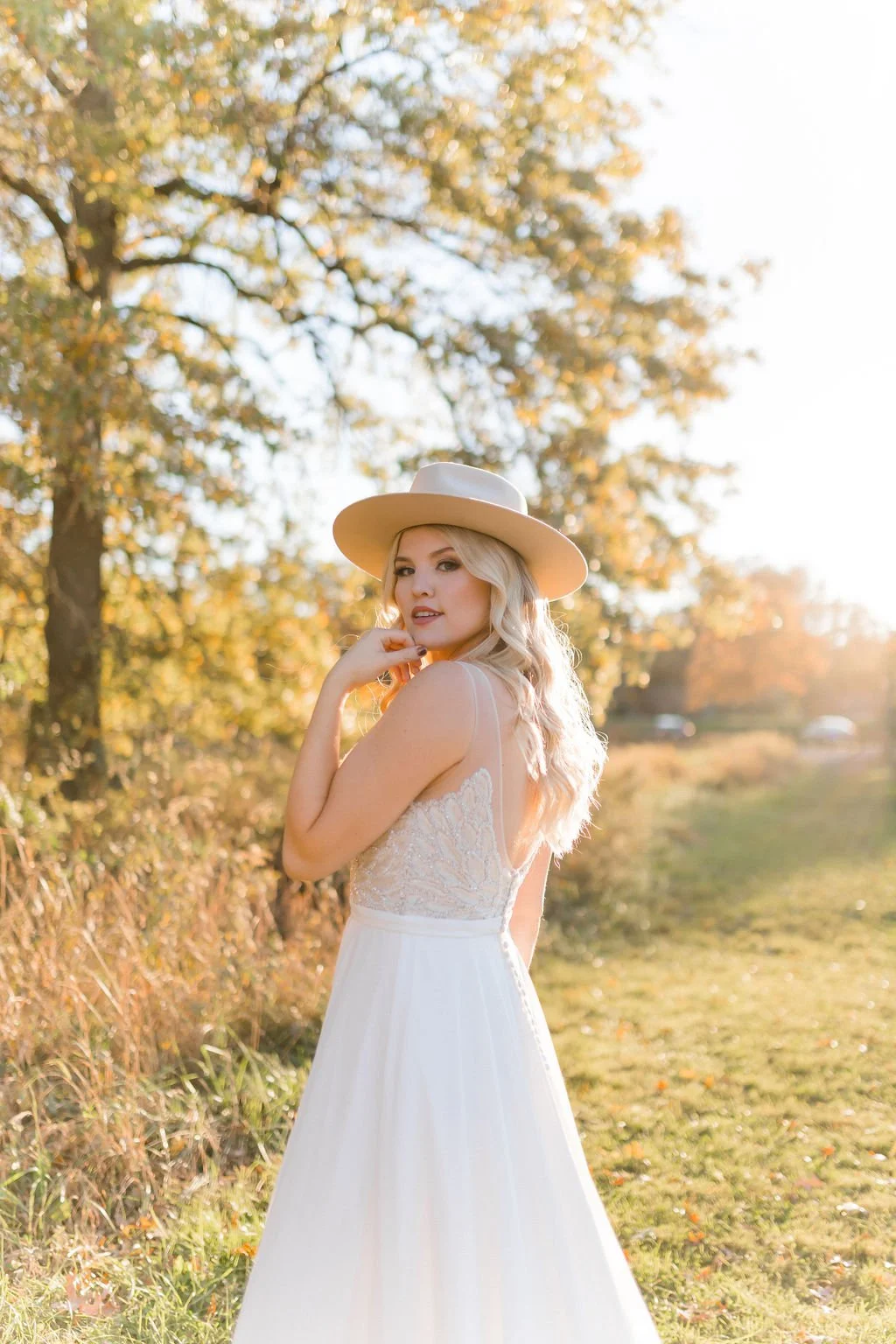 A young woman in a white lace dress and beige wide-brimmed hat standing outdoors during late afternoon, with trees and sunlight in the background.