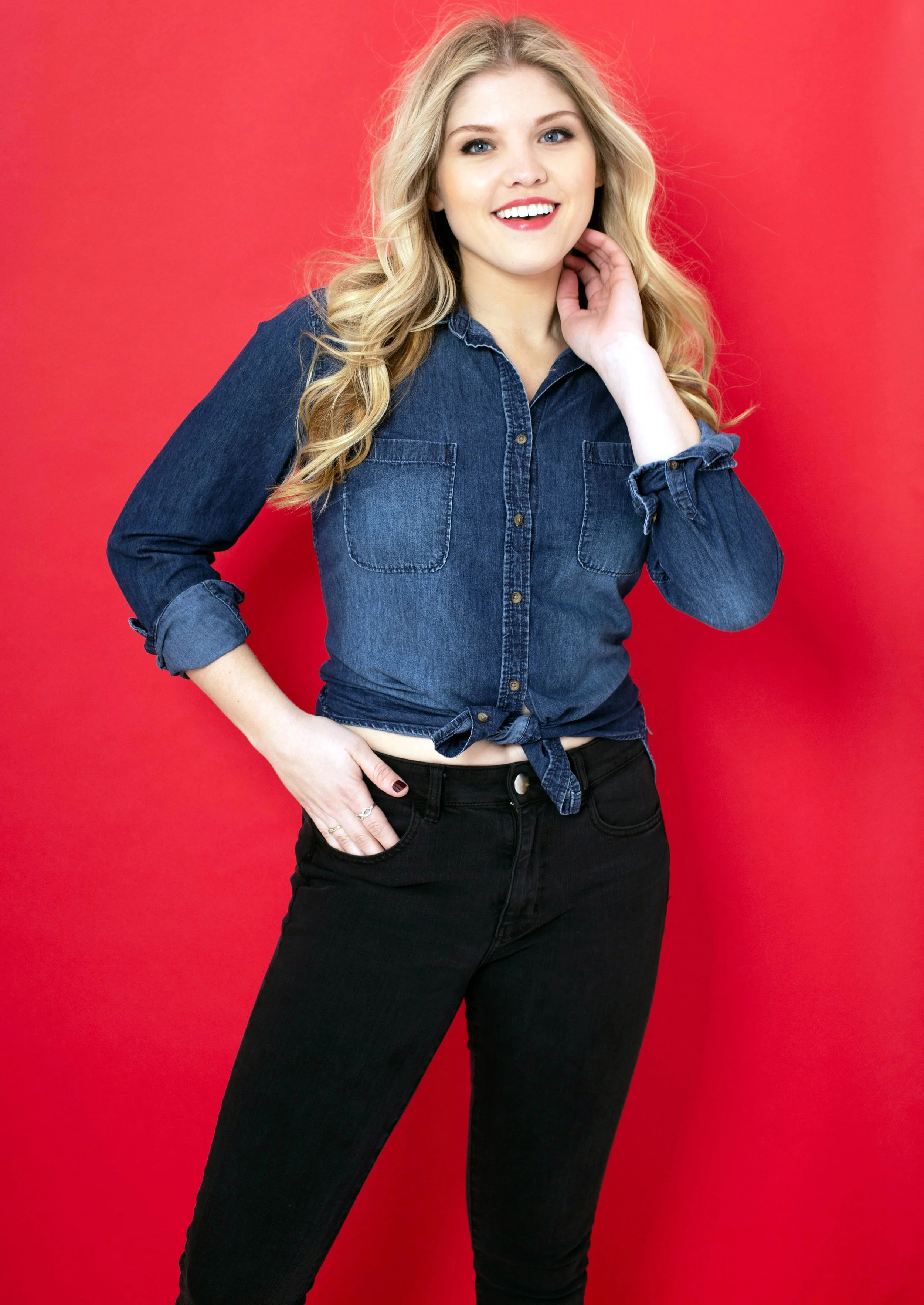 Young blonde woman smiling, wearing a denim shirt and black jeans, standing against a red background.