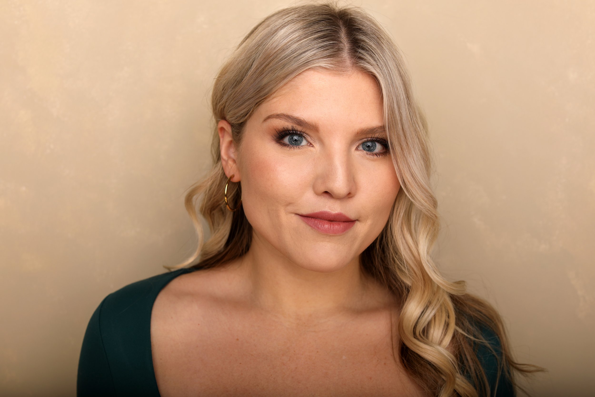 Close-up portrait of a woman with long wavy blonde hair, blue eyes, and gold hoop earrings, smiling softly against a beige background.