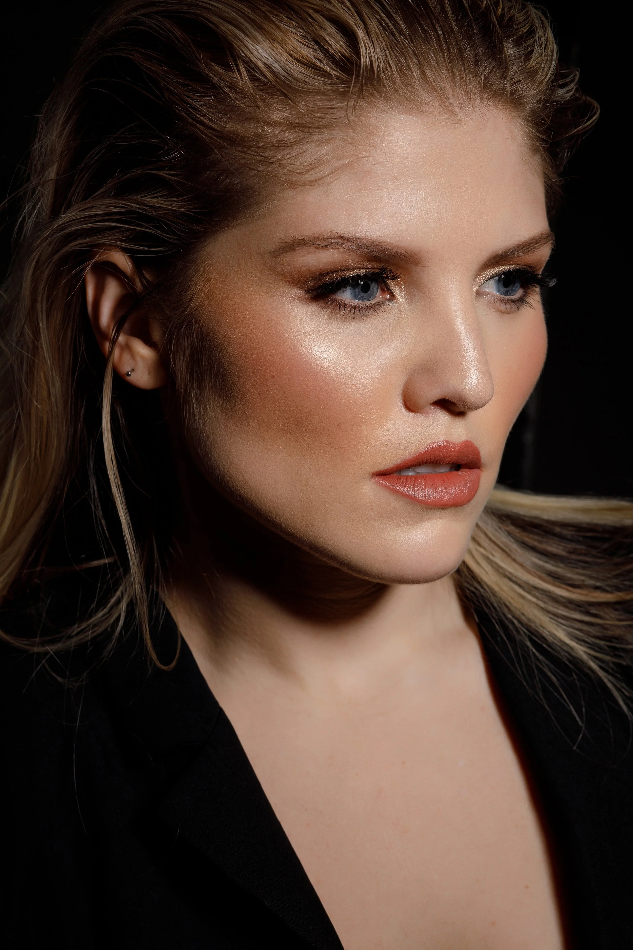 Close-up of a woman's face with fair skin, blue eyes, light makeup, and wavy blonde hair, wearing a black top against a black background.