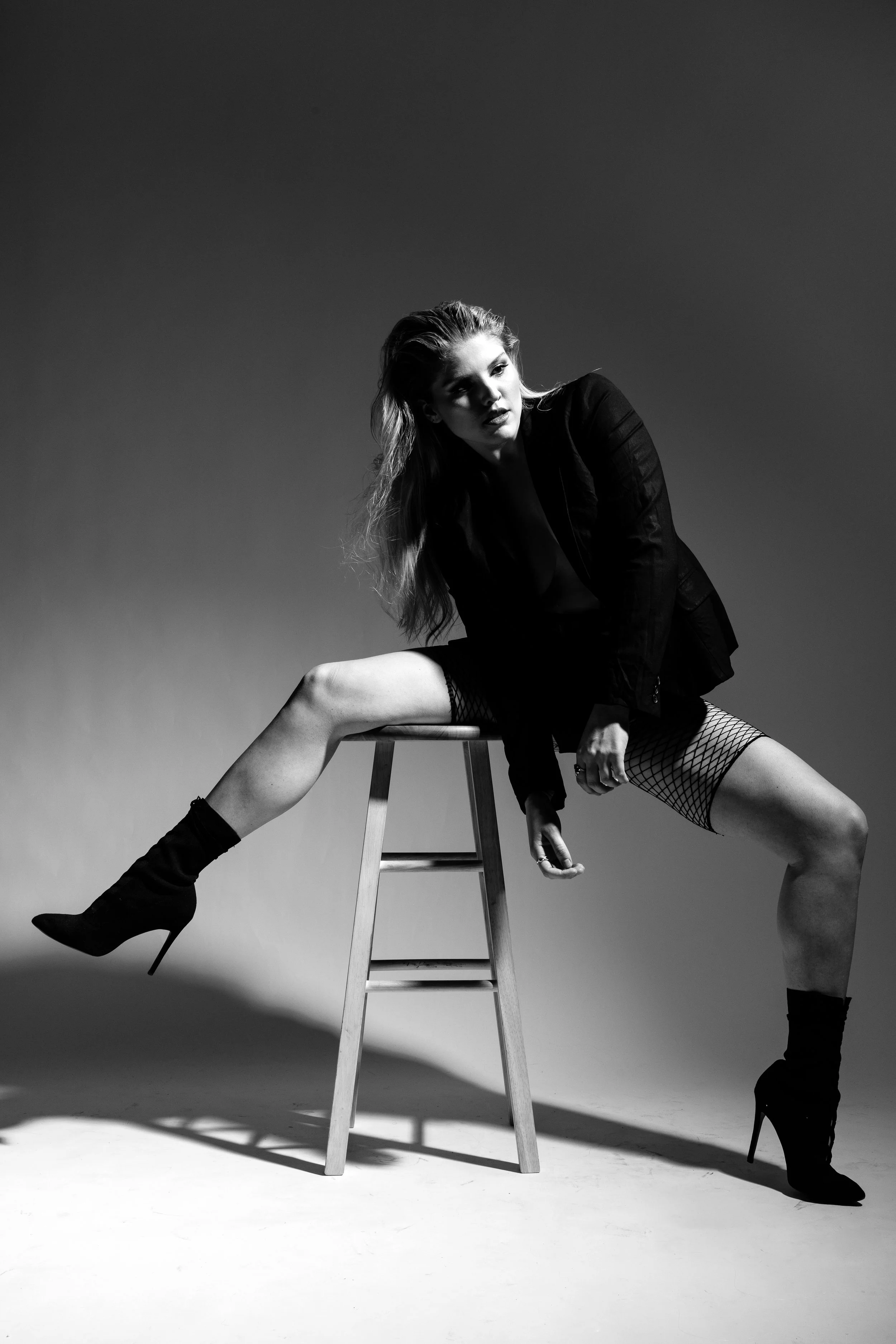 Black and white photo of a woman with wavy hair, sitting on a wooden stool with legs spread apart. She is wearing high-heeled boots, fishnet stockings, and a black jacket. One arm rests on her knee while the other hand touches her thigh. The backgrou