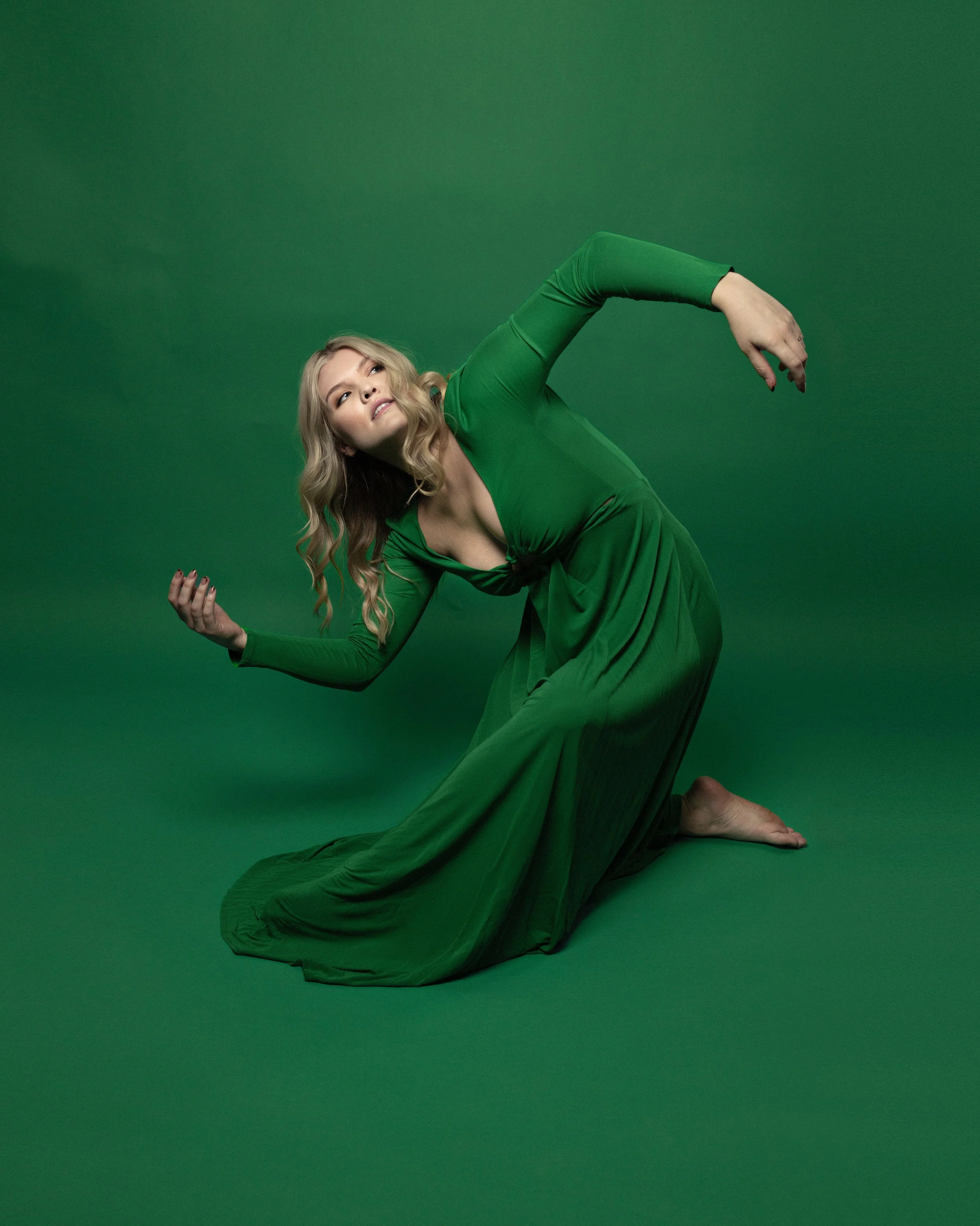 A woman in a green dress posed on a green background, kneeling with one arm raised and the other hand extended, with her head tilted and looking to the side.