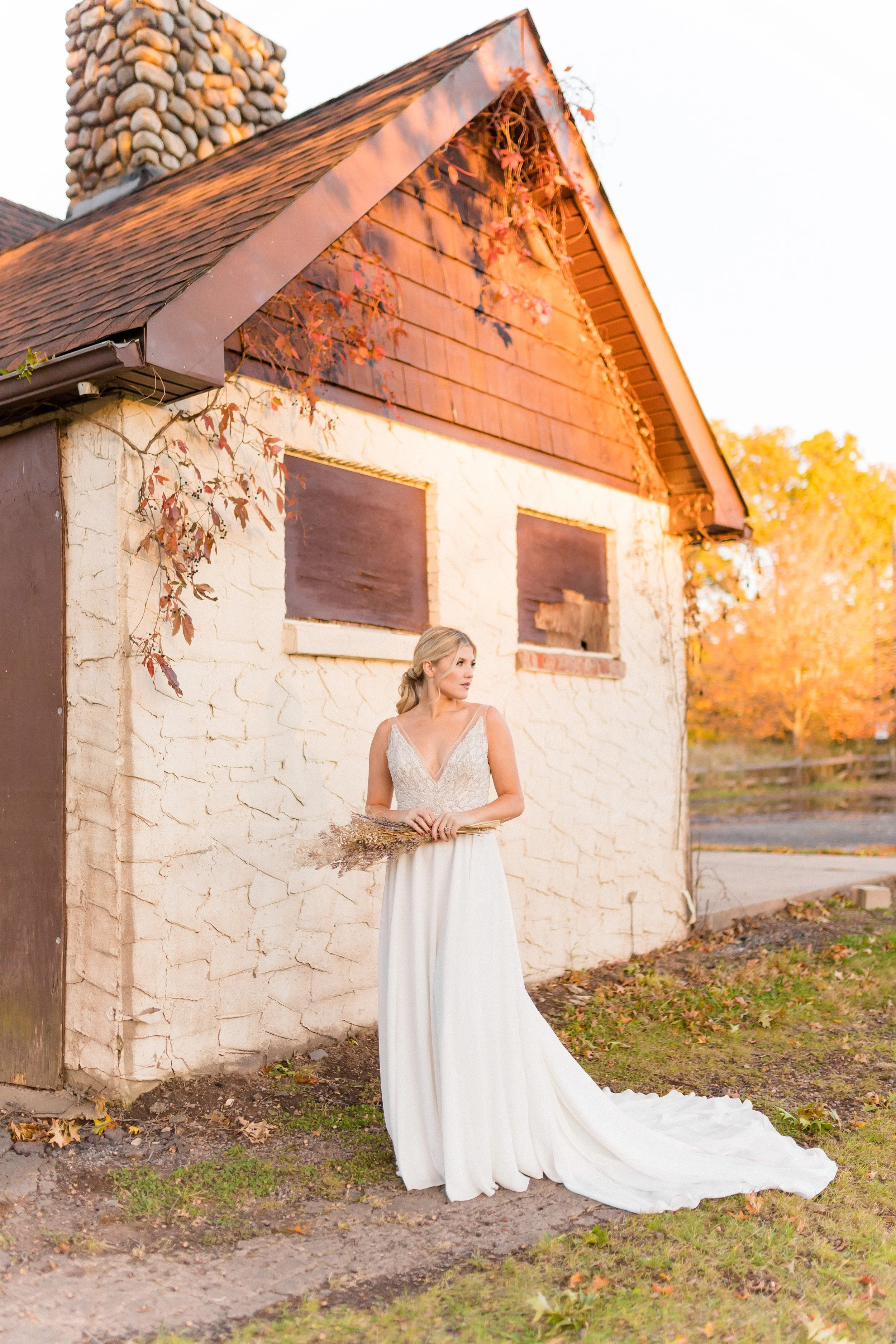 A bride standing outdoors next to a rustic stone and stucco building with autumn foliage in the background. She is wearing a white wedding dress with lace details and holding a bouquet of dried flowers, looking to her right.