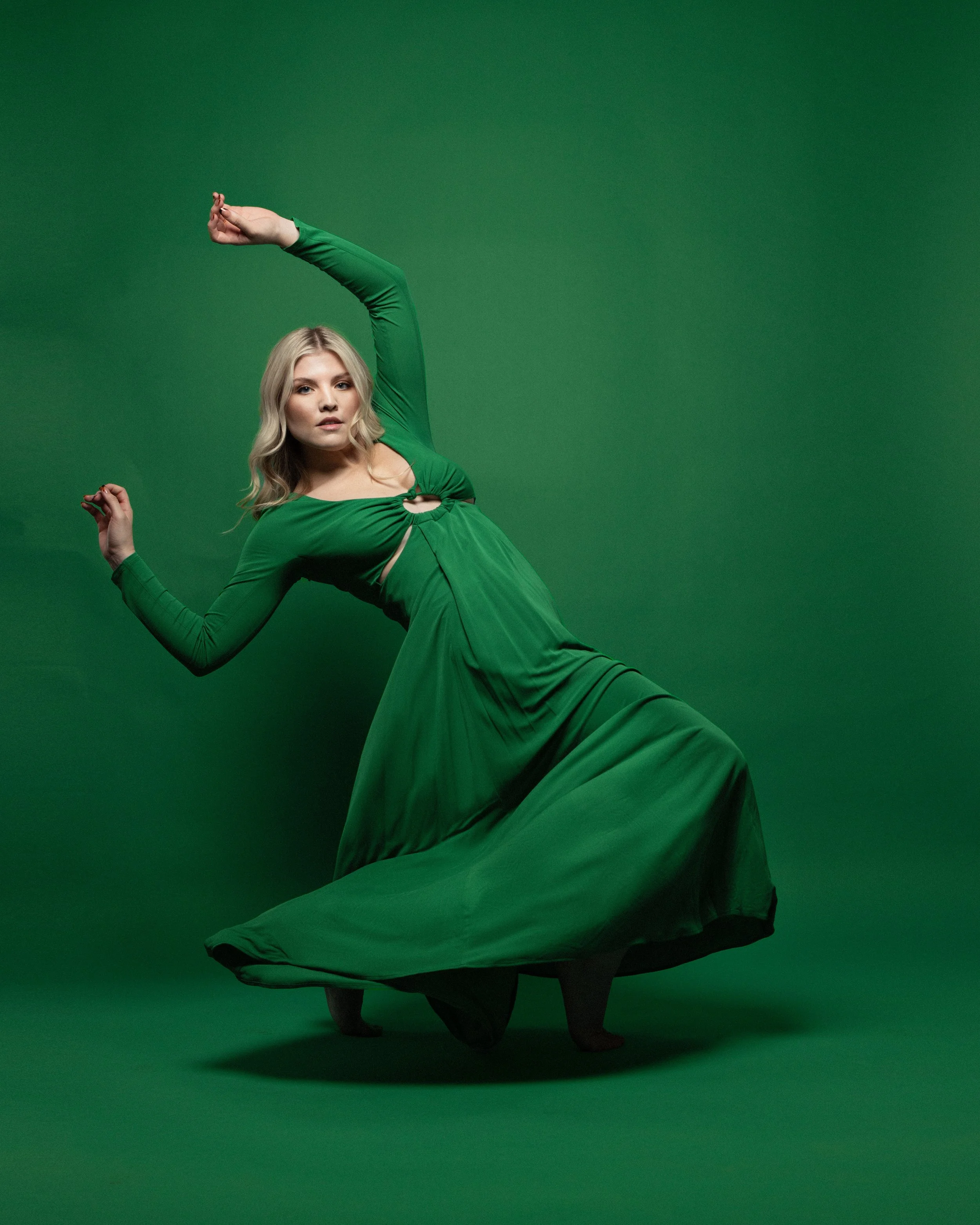 A woman in a long green dress, standing against a green background, poses with one arm arched above her head and the other bent at her side.