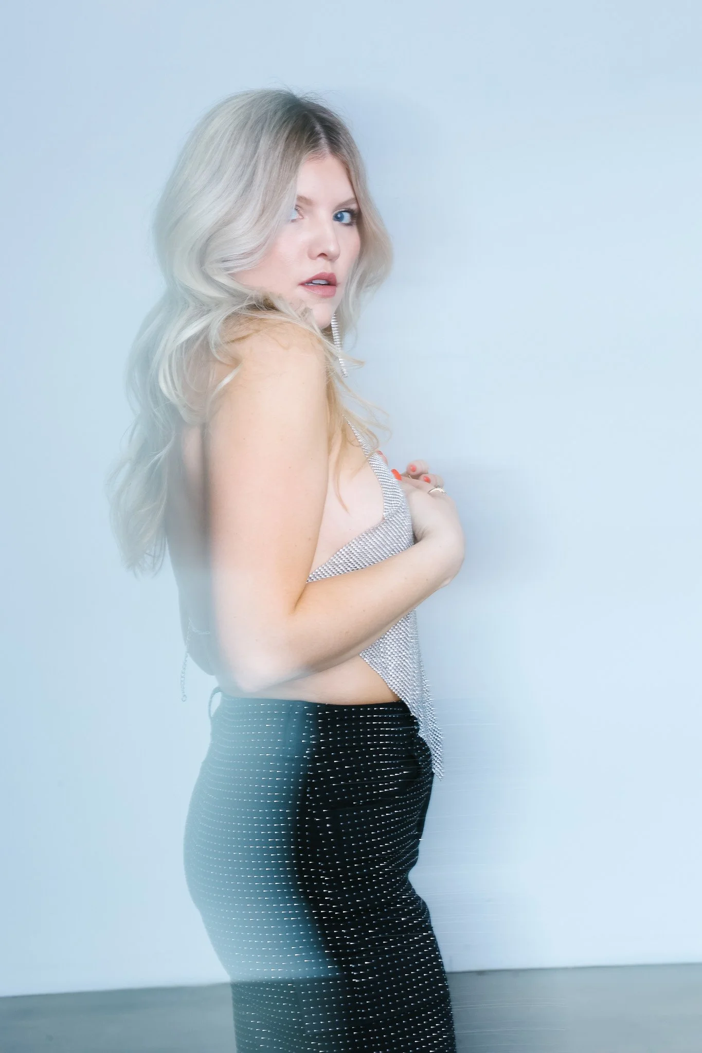 A woman with long blonde hair, wearing a silver top and black pants with white polka dots, standing against a plain gray wall.