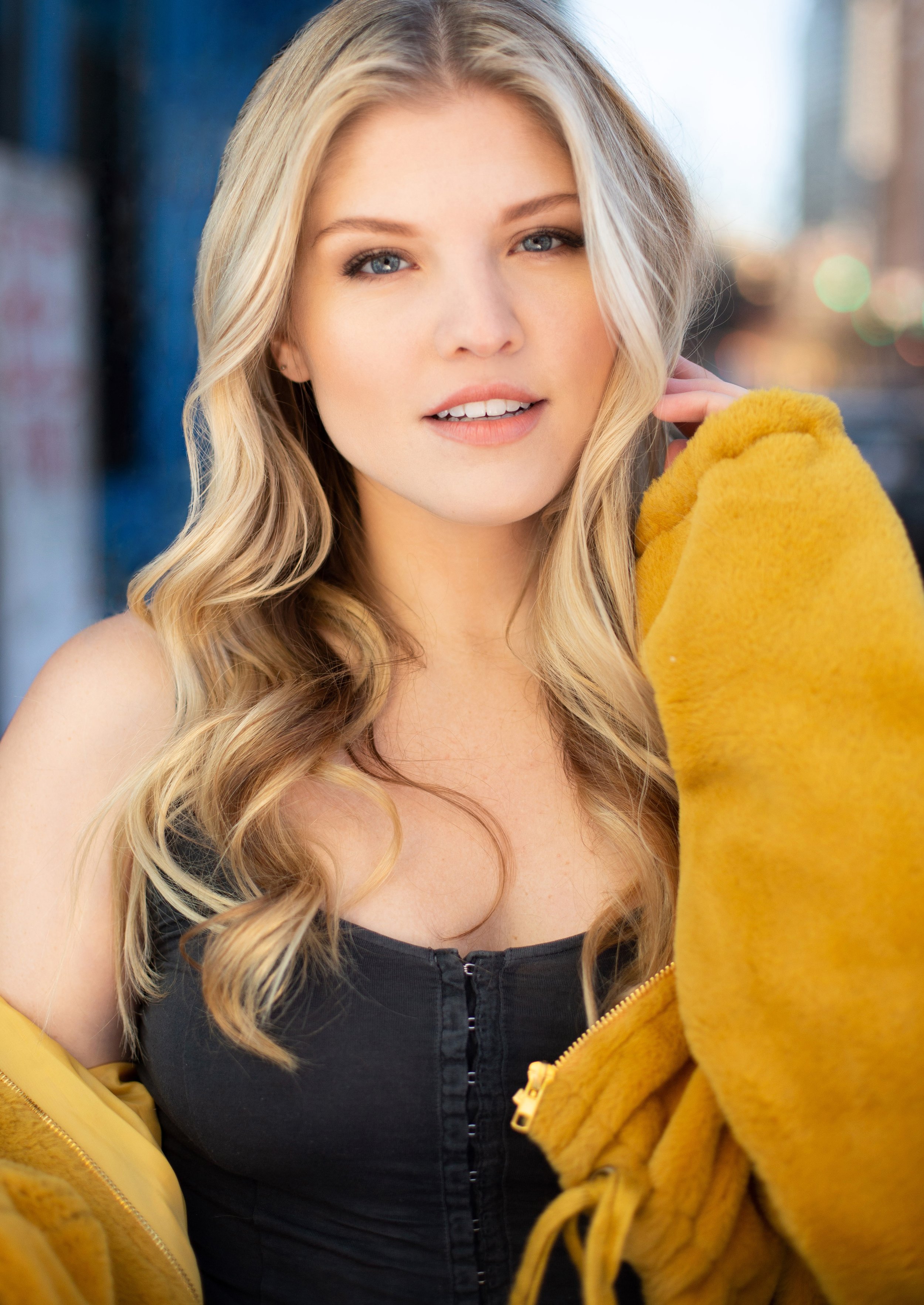 Close-up of a young woman with long, wavy blonde hair and blue eyes, wearing a yellow jacket and a black top, standing outdoors in an urban setting with city lights in the background.