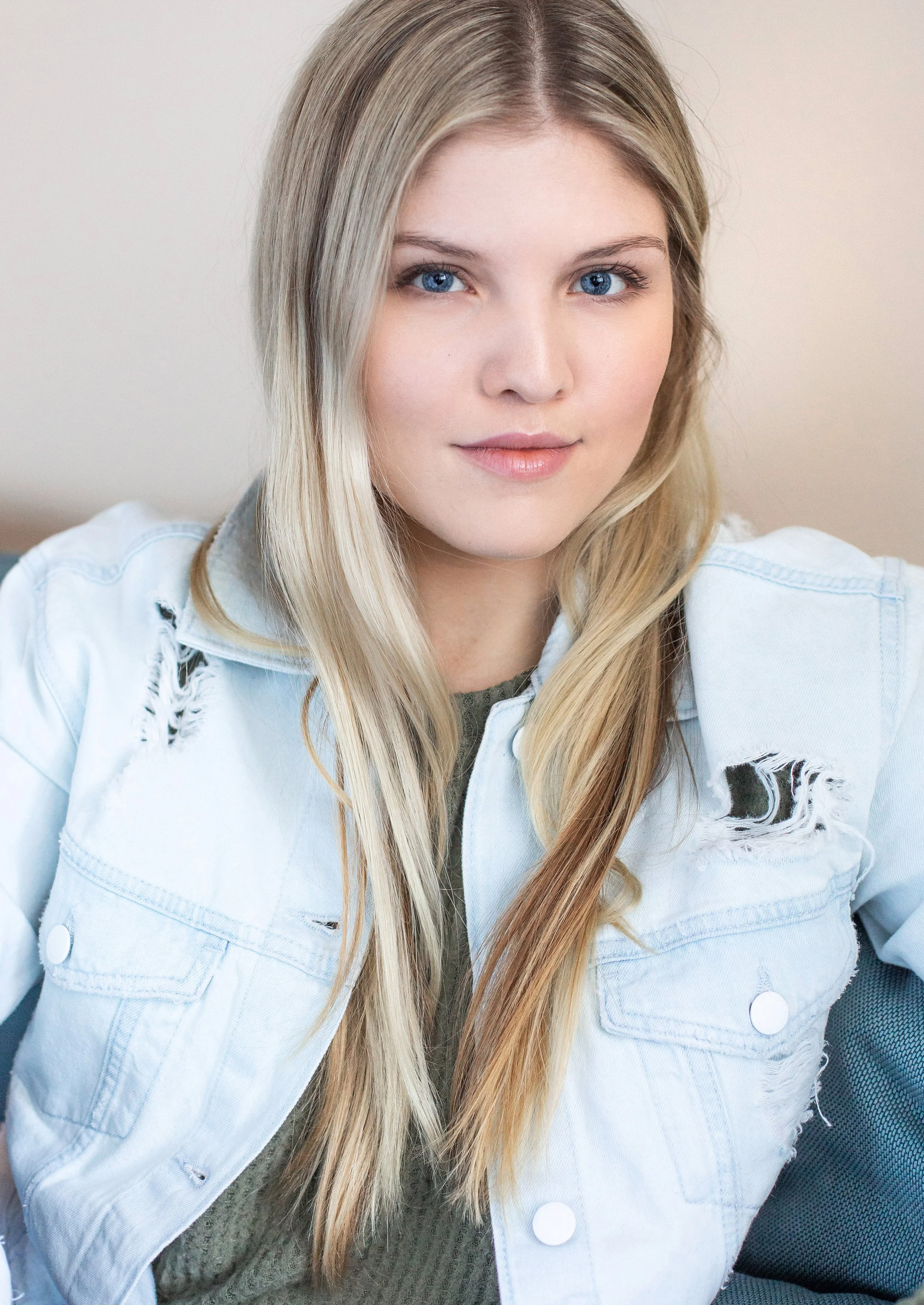 A young woman with long blonde hair and blue eyes wearing a distressed white denim jacket and a green sweater, sitting in a car.