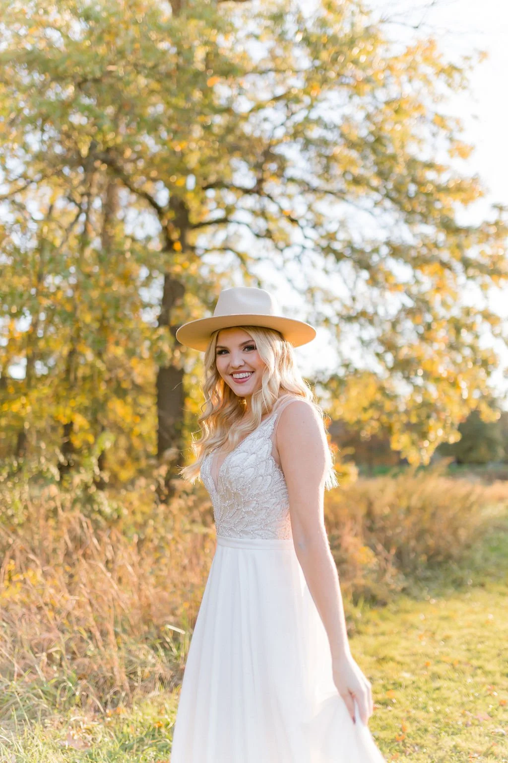 A woman with blonde wavy hair smiling and wearing a white dress and a cream-colored wide-brimmed hat, standing outdoors in a field with trees showing fall foliage.