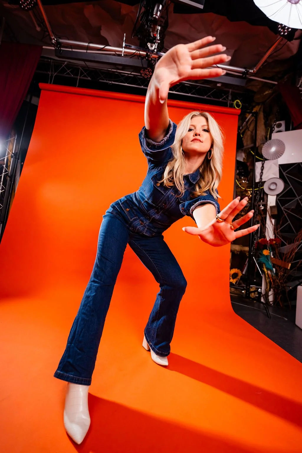 A woman in denim jeans and a denim jacket crouches with arms extended towards the camera, standing on an orange backdrop in a photography studio.