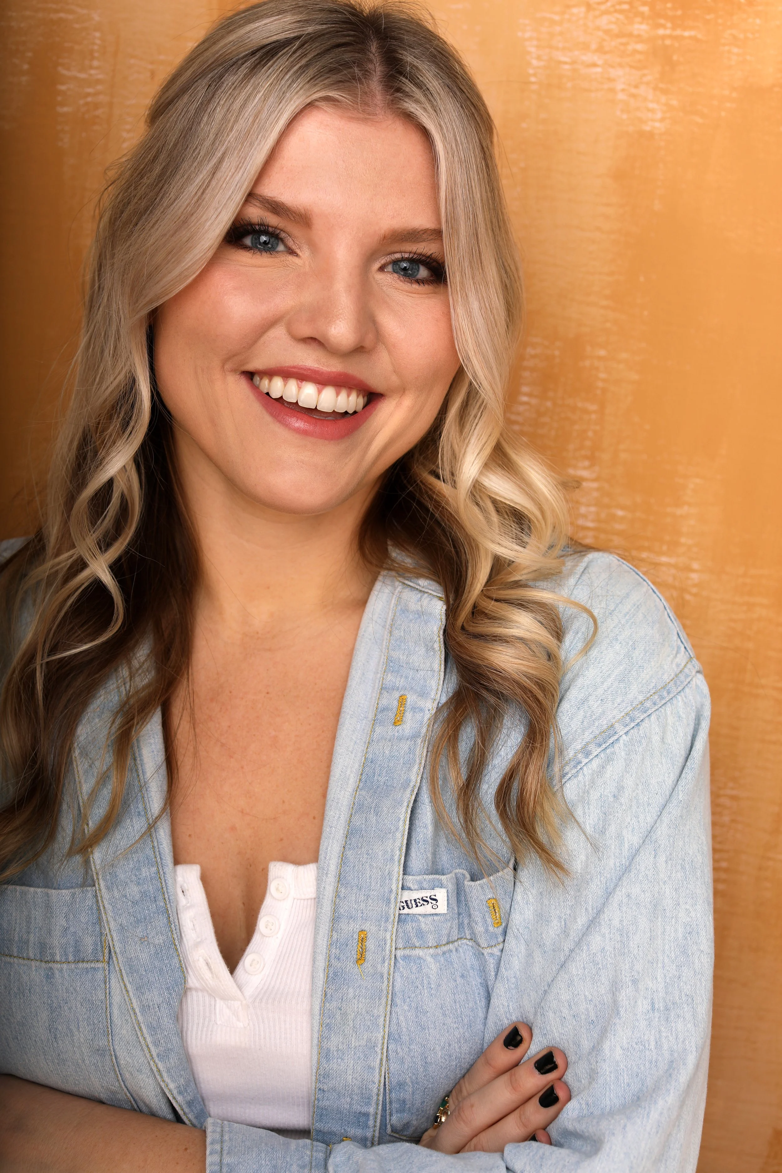 A young woman with blonde, wavy hair smiling, wearing a layered denim jacket and white top, against a wooden background.