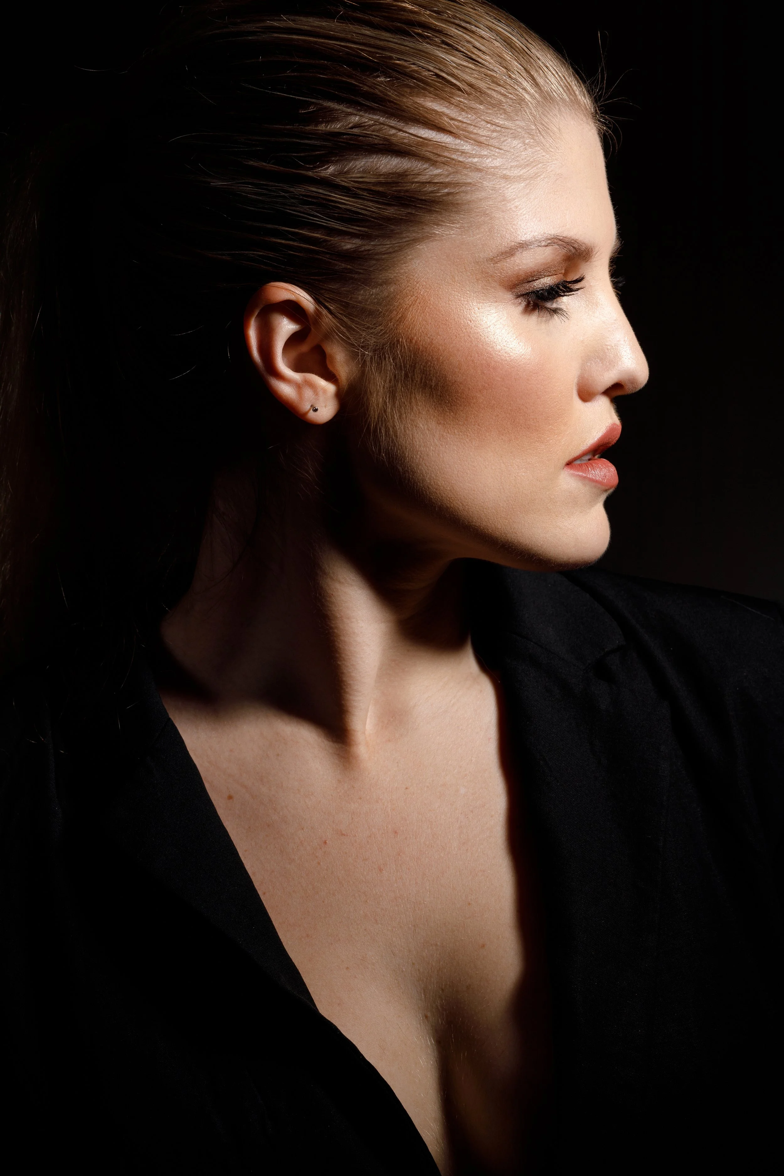 Profile of a woman with light skin and brown hair tied back, wearing subtle makeup and a black top, against a black background.