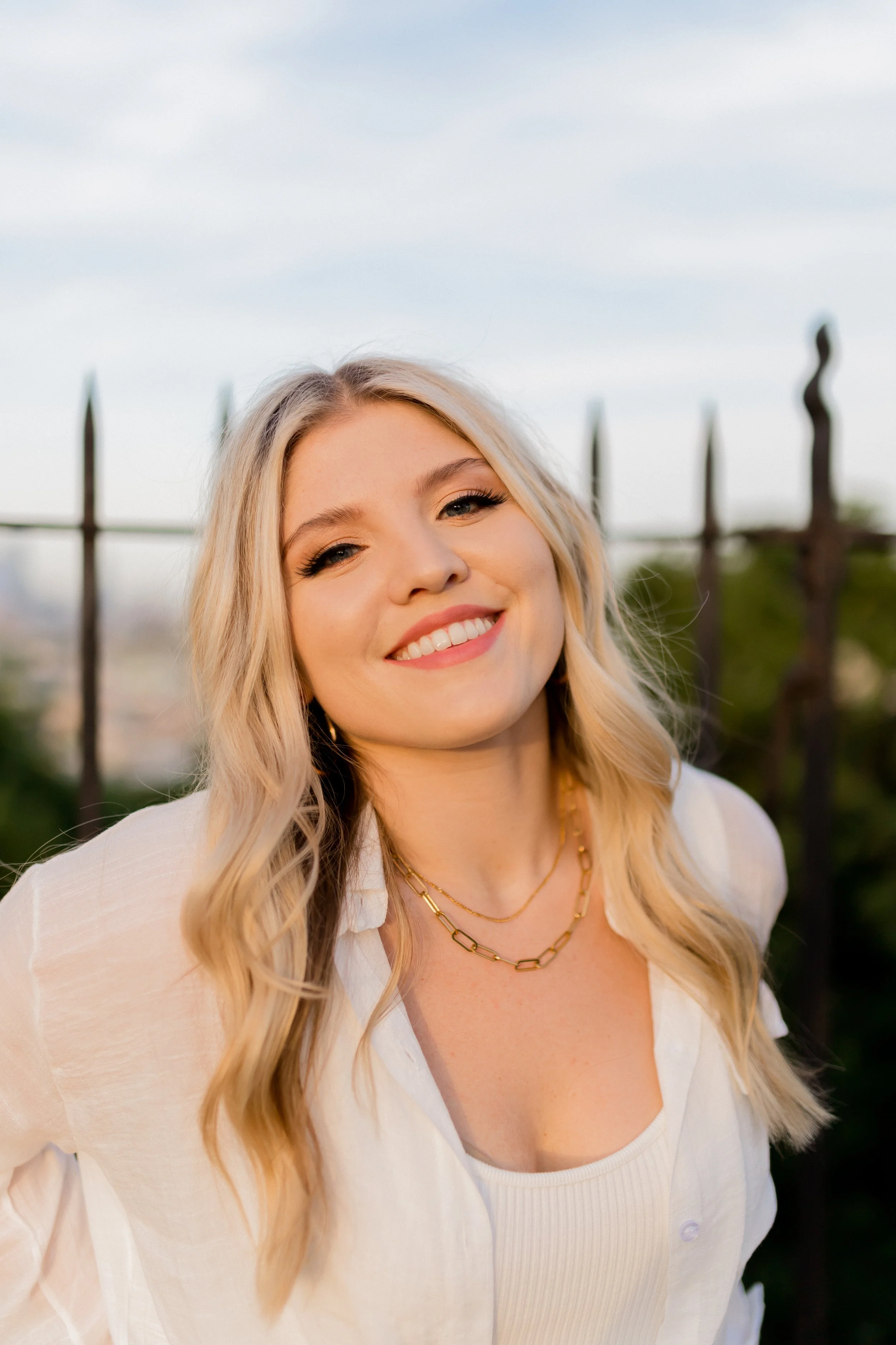 A young woman with blonde hair styled in loose waves, wearing a white shirt, gold layered necklaces, and smiling outdoors with a blurred background of a fence and trees.