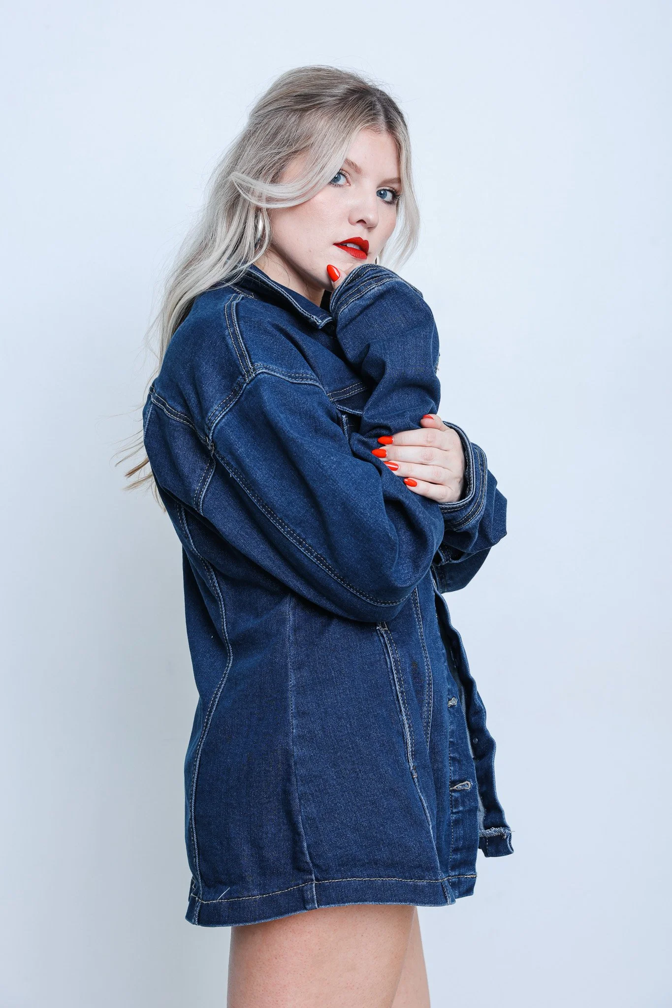 A woman with platinum blonde hair, red lipstick, and blue eyes wearing a dark blue denim jacket with her hand touching her face. The background is plain and light-colored.