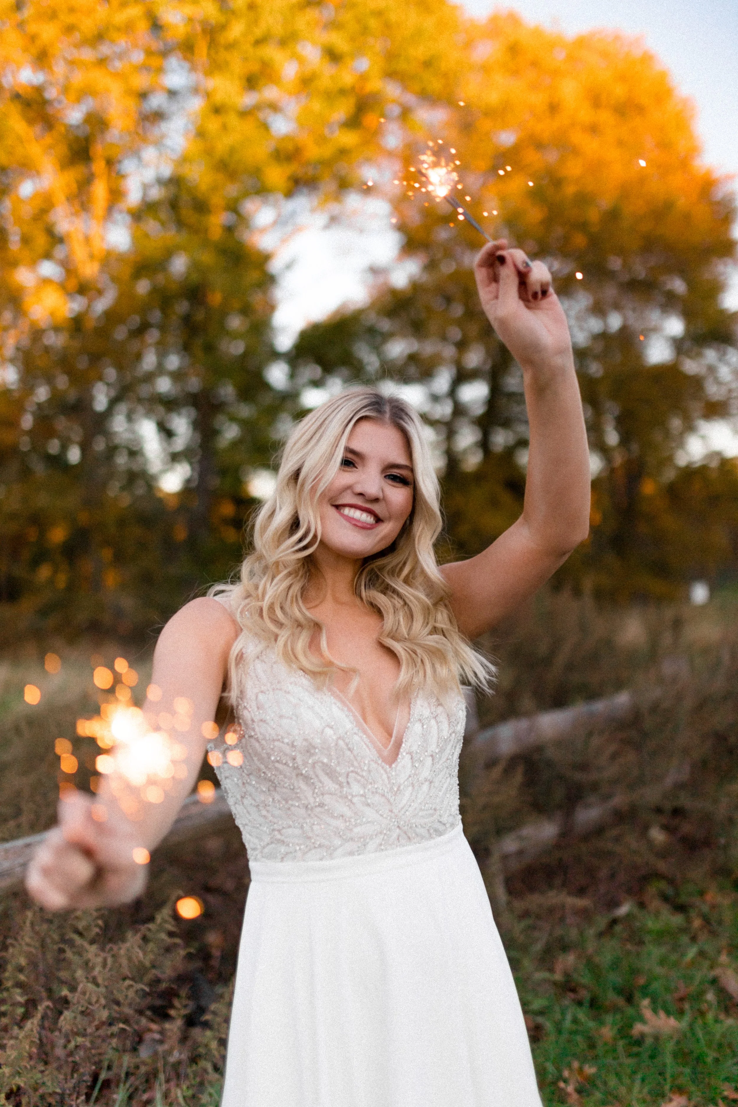 A smiling young woman in a white dress holding a sparkler outdoors during sunset with trees in the background.