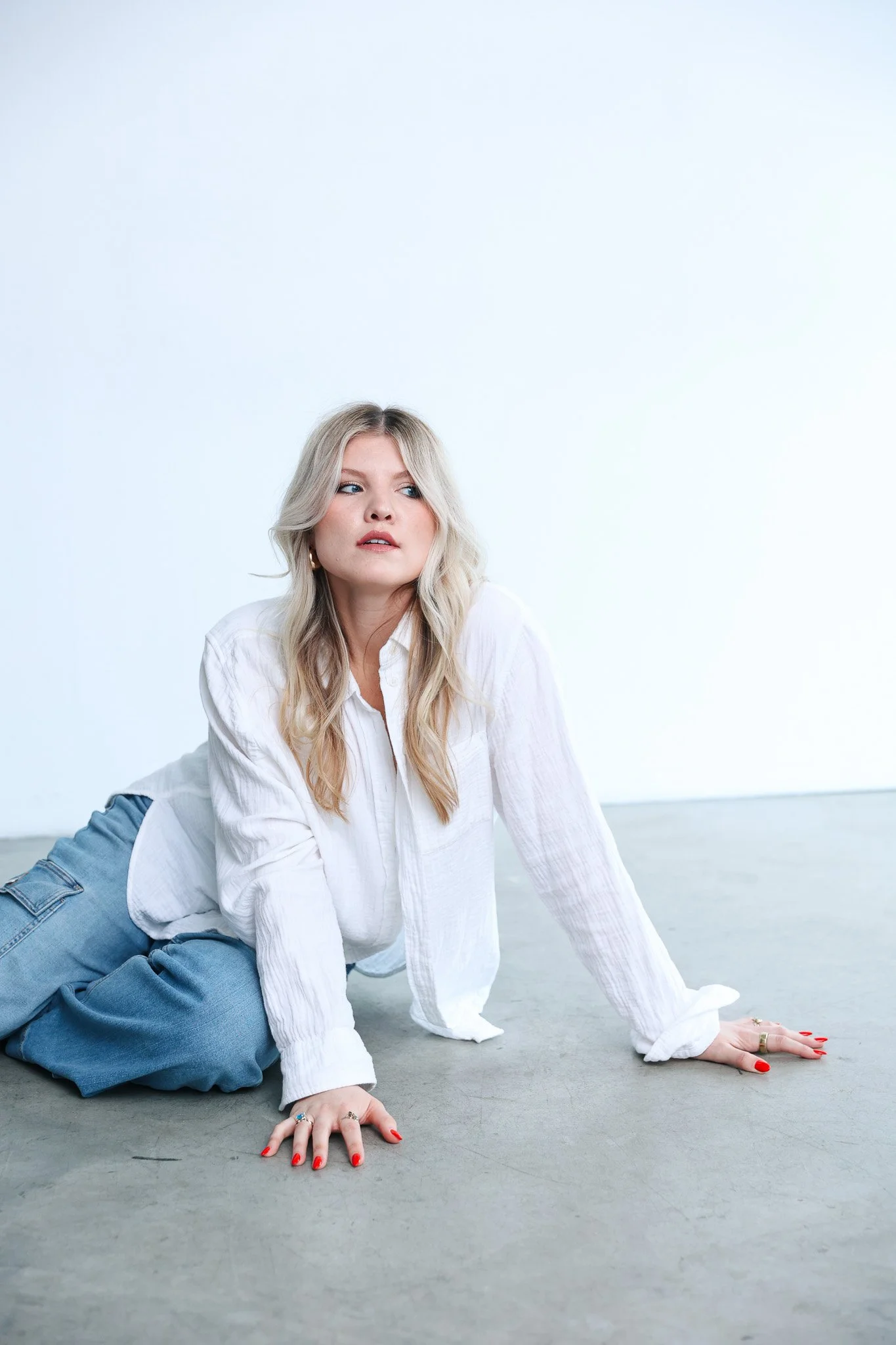 A woman with long blonde hair is posing on the floor, supporting herself with her hands and knees, in a white room with no visible furniture or decorations.