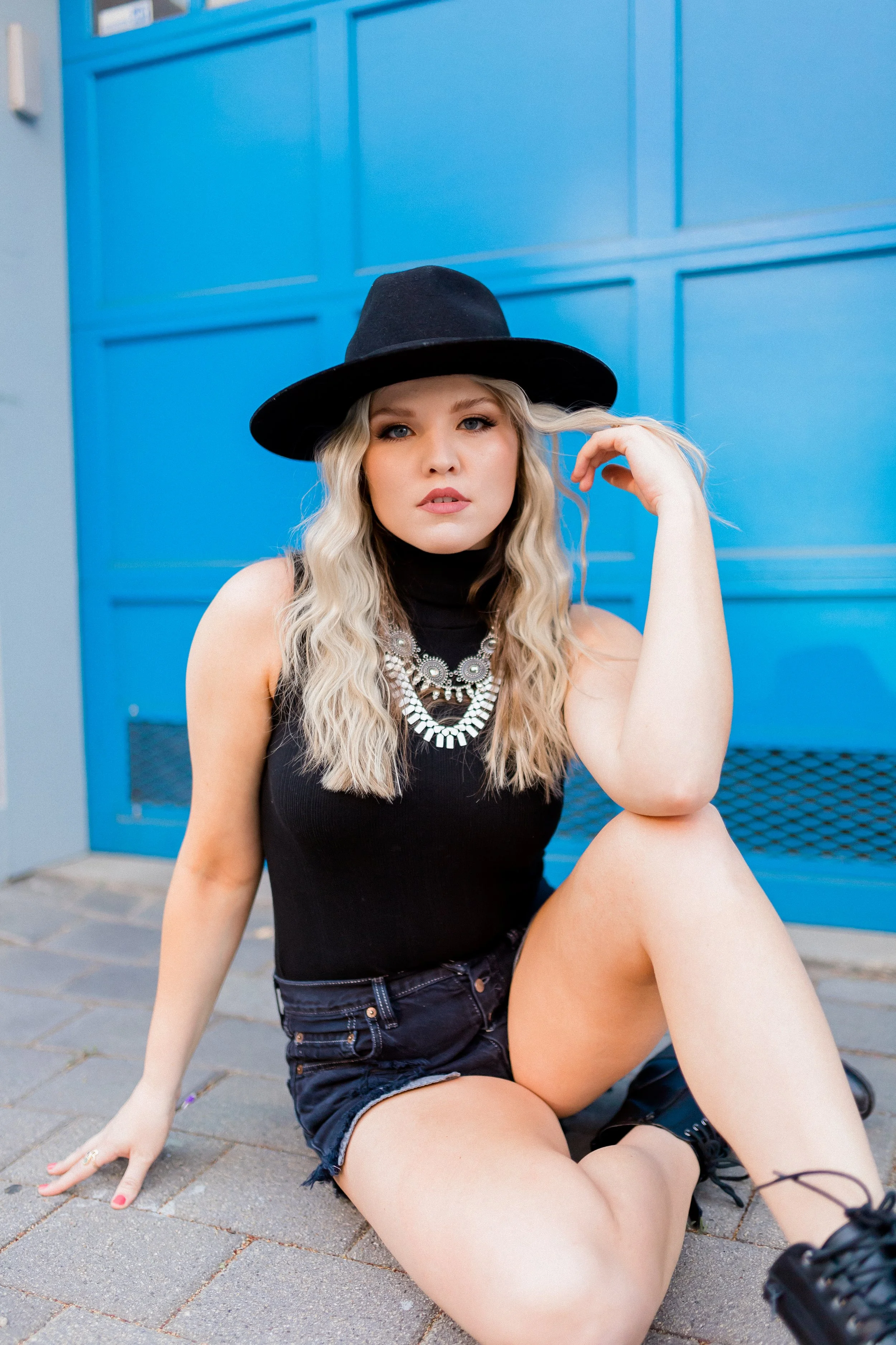 A blonde woman wearing a black wide-brimmed hat, black sleeveless top, and jewelry, sitting on the ground in front of a blue garage door, with one leg bent and her hand resting on her knee.