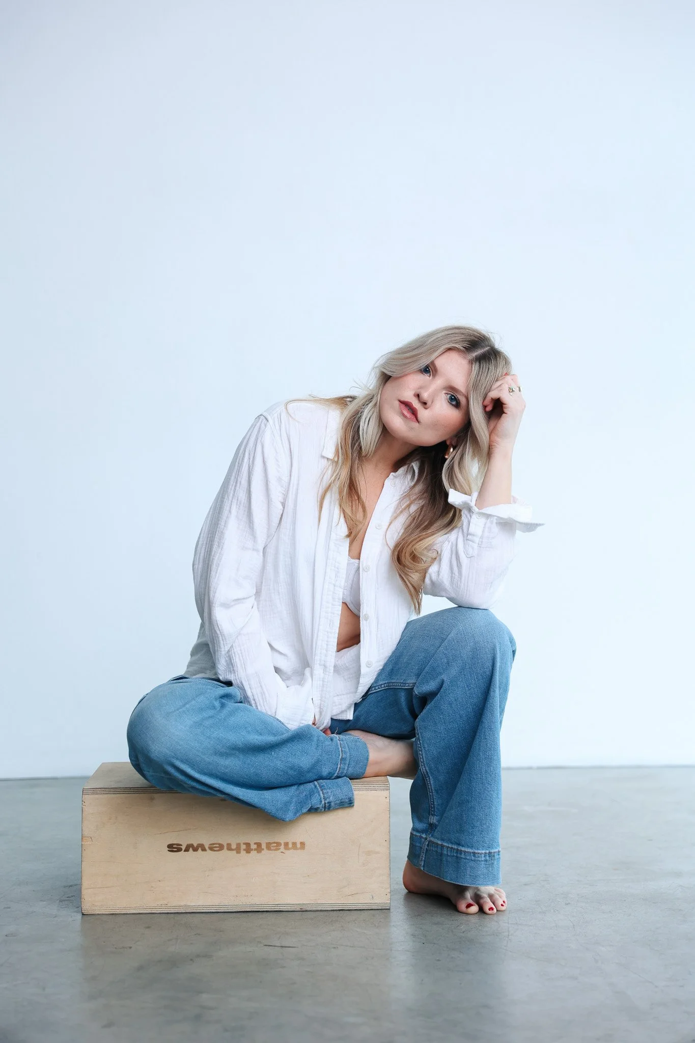 A woman with blonde wavy hair and blue eyes sitting on a wooden box with the word "mallthings" on it, wearing a loose white shirt and blue jeans, with her hand resting on her head, looking relaxed against a minimalist light blue background.