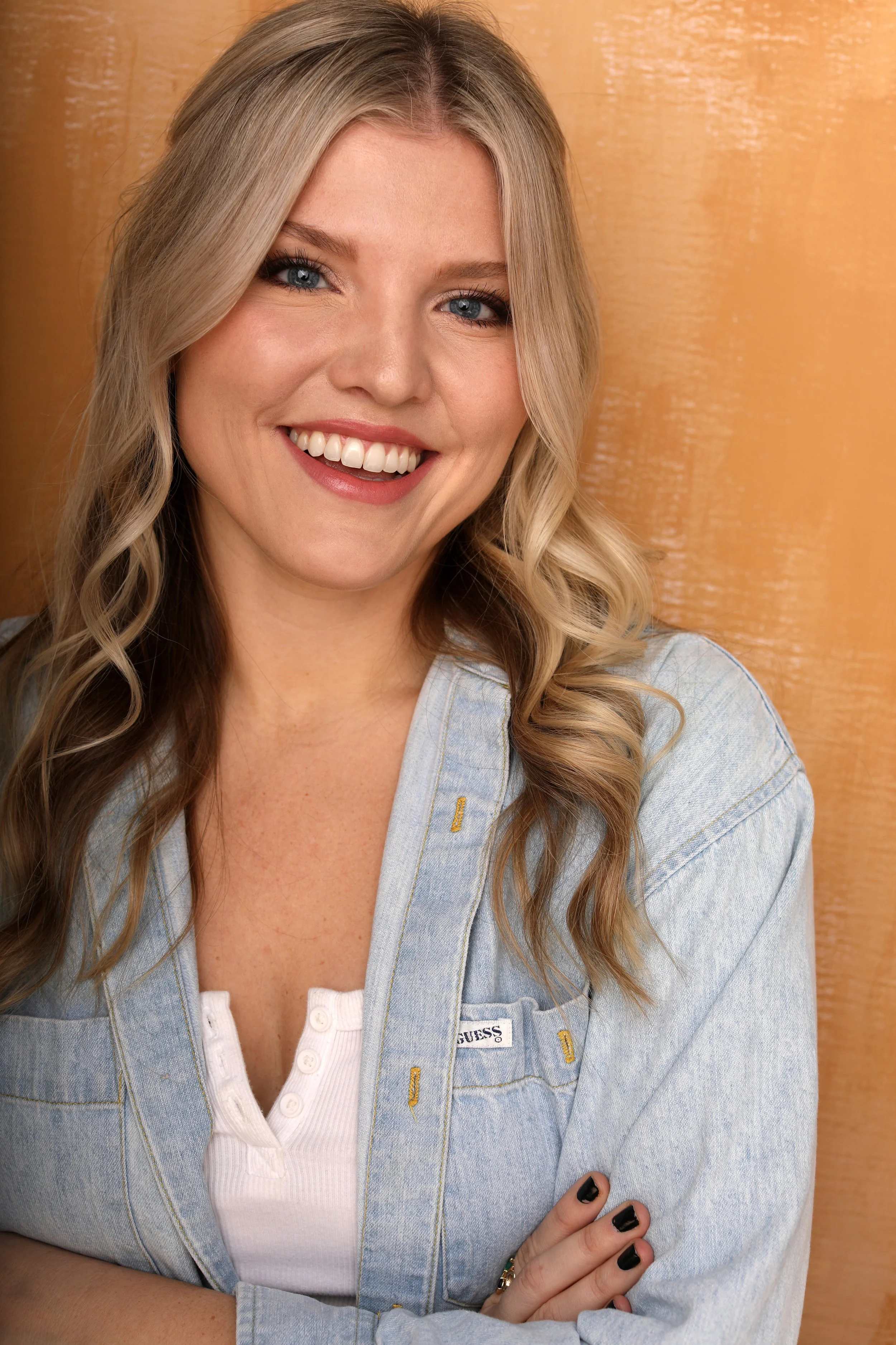 Close-up of a smiling woman with long, wavy blonde hair wearing a light blue denim jacket over a white top, standing in front of a wooden background.
