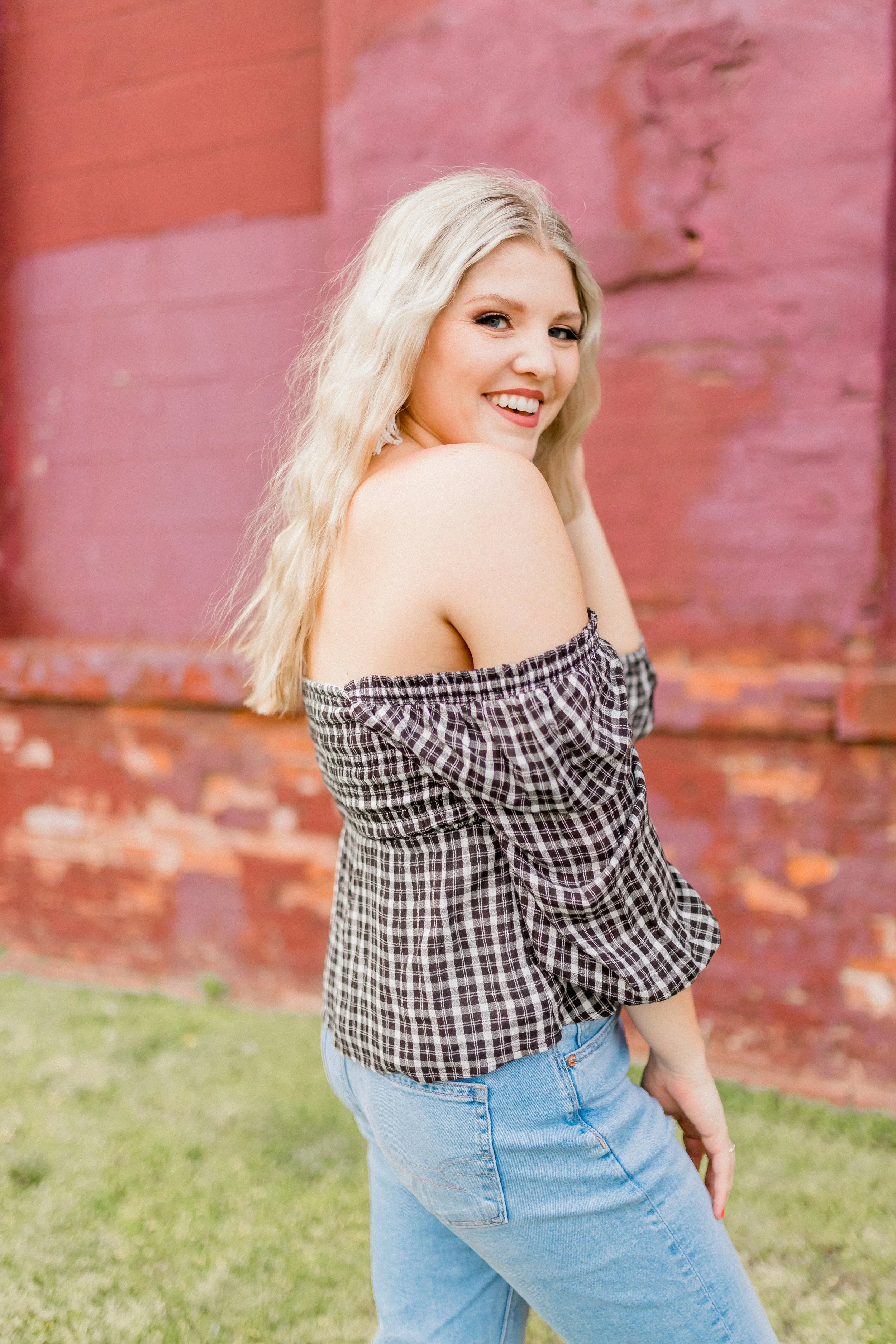 A smiling young woman with long blonde hair, wearing a black and white checkered off-shoulder top and blue jeans, standing in front of a pink brick wall outdoors.